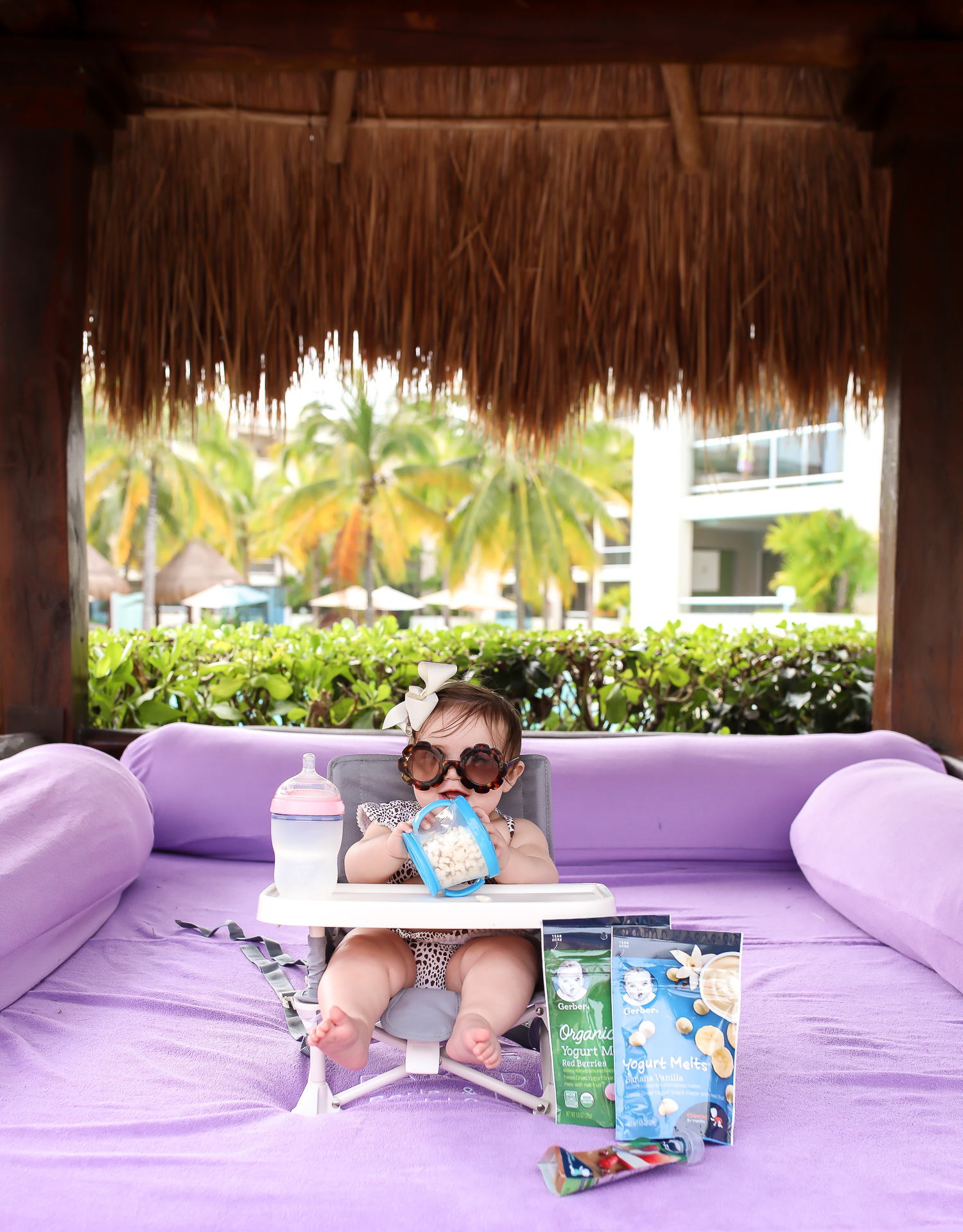 Christmas In Cancun🌟🎄🎁 [+ A Few Baby Travel Must-Haves] by popular Oklahoma travel blog, The Sweetest Thing: image of a baby in Cancun, Mexico sitting in a Walmart hiccapop Omniboost Travel Booster Seat with Tray for Baby holding a Walmart Munchkin Snack Catcher Snack Cup with Gerber Puff cereal snack inside, with a bottle resting on her high chair tray and some Gerber Organic Yogurt melts next to her high chair. 