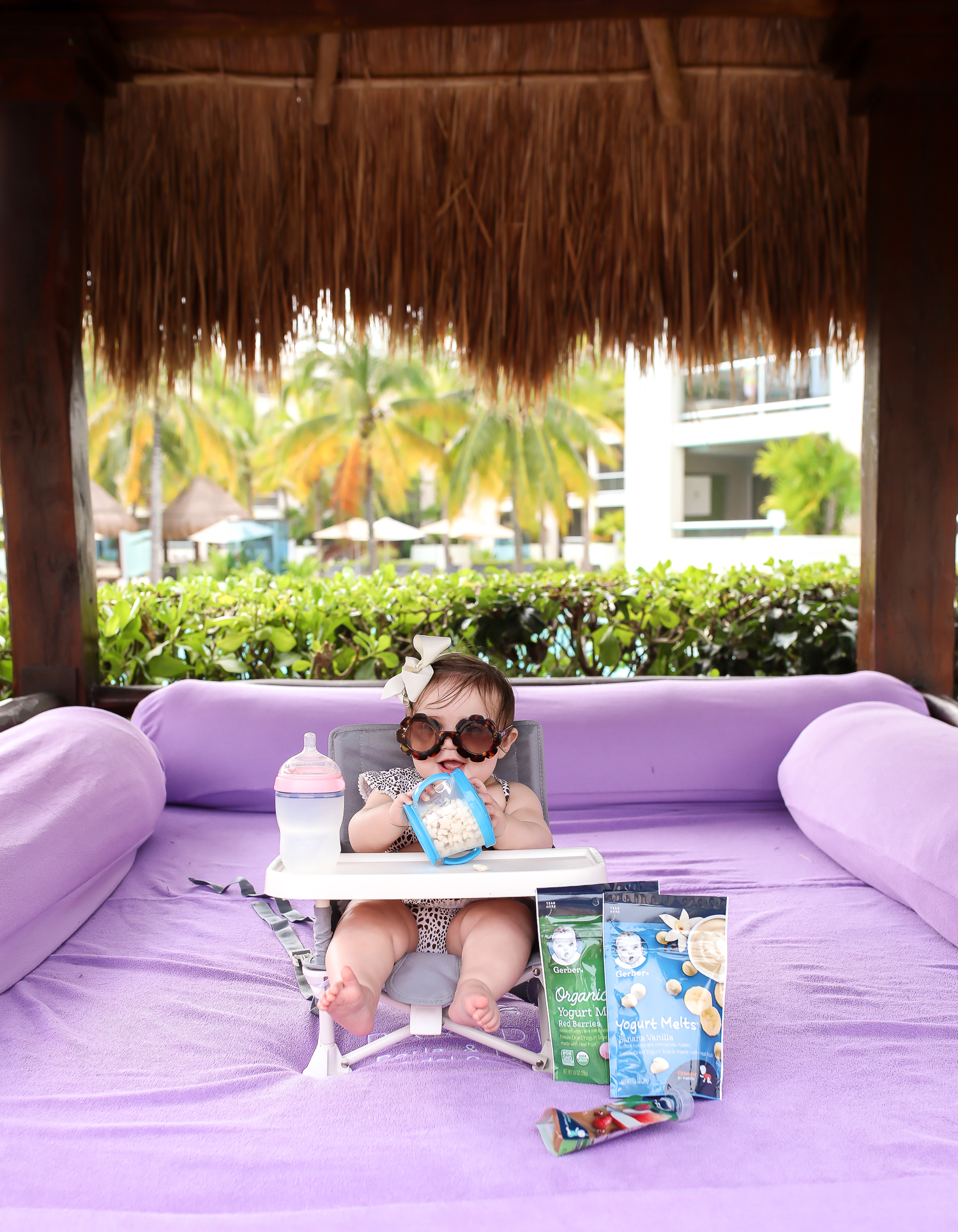 Christmas In Cancun🌟🎄🎁 [+ A Few Baby Travel Must-Haves] by popular Oklahoma travel blog, The Sweetest Thing: image of a baby in Cancun, Mexico sitting in a Walmart hiccapop Omniboost Travel Booster Seat with Tray for Baby holding a Walmart Munchkin Snack Catcher Snack Cup with Gerber Puff cereal snack inside, with a bottle resting on her high chair tray and some Gerber Organic Yogurt melts next to her high chair. 