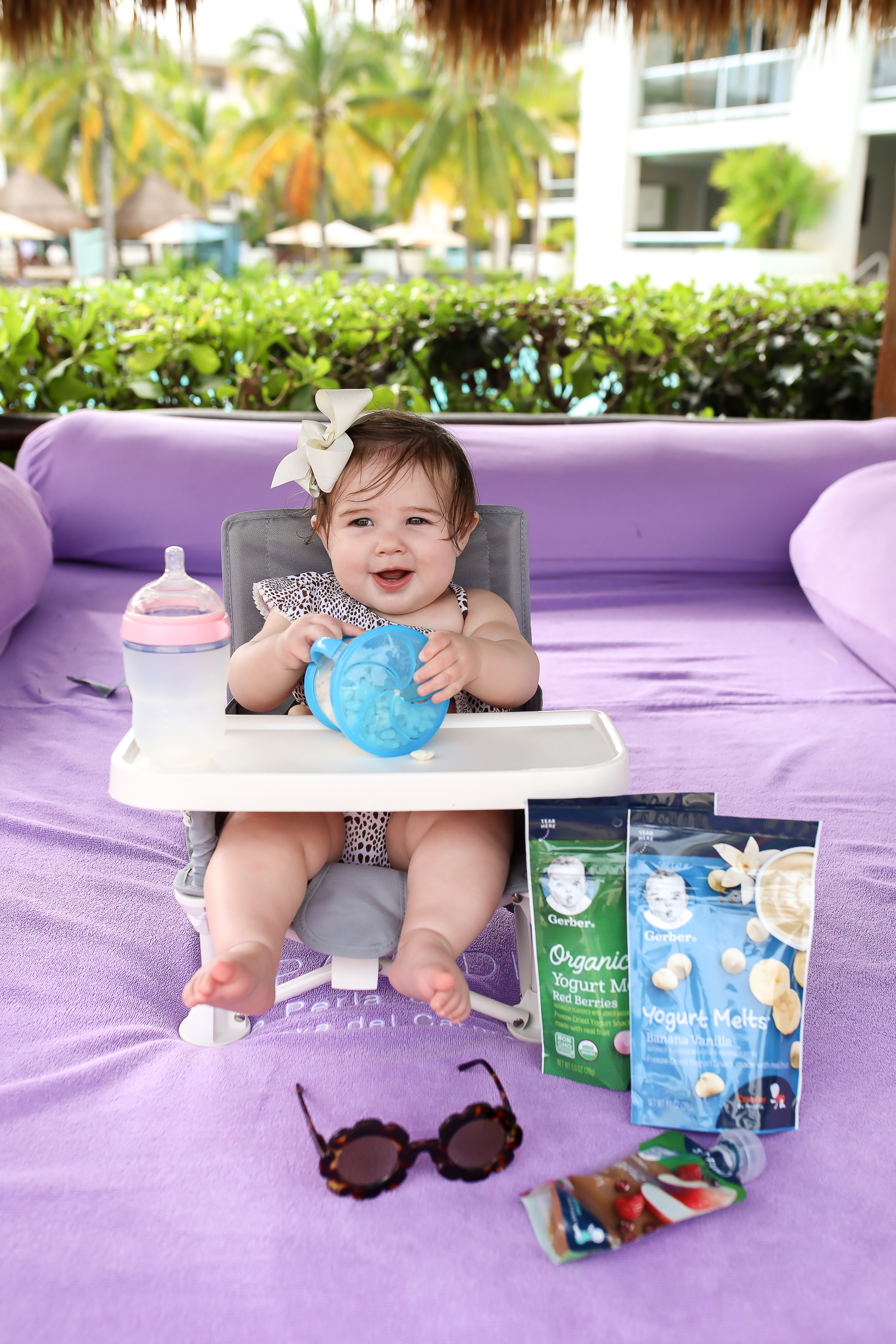 Christmas In Cancun🌟🎄🎁 [+ A Few Baby Travel Must-Haves] by popular Oklahoma travel blog, The Sweetest Thing: image of a baby in Cancun, Mexico sitting in a Walmart hiccapop Omniboost Travel Booster Seat with Tray for Baby holding a Walmart Munchkin Snack Catcher Snack Cup with Gerber Puff cereal snack inside, with a bottle resting on her high chair tray and some Gerber Organic Yogurt melts next to her high chair. 