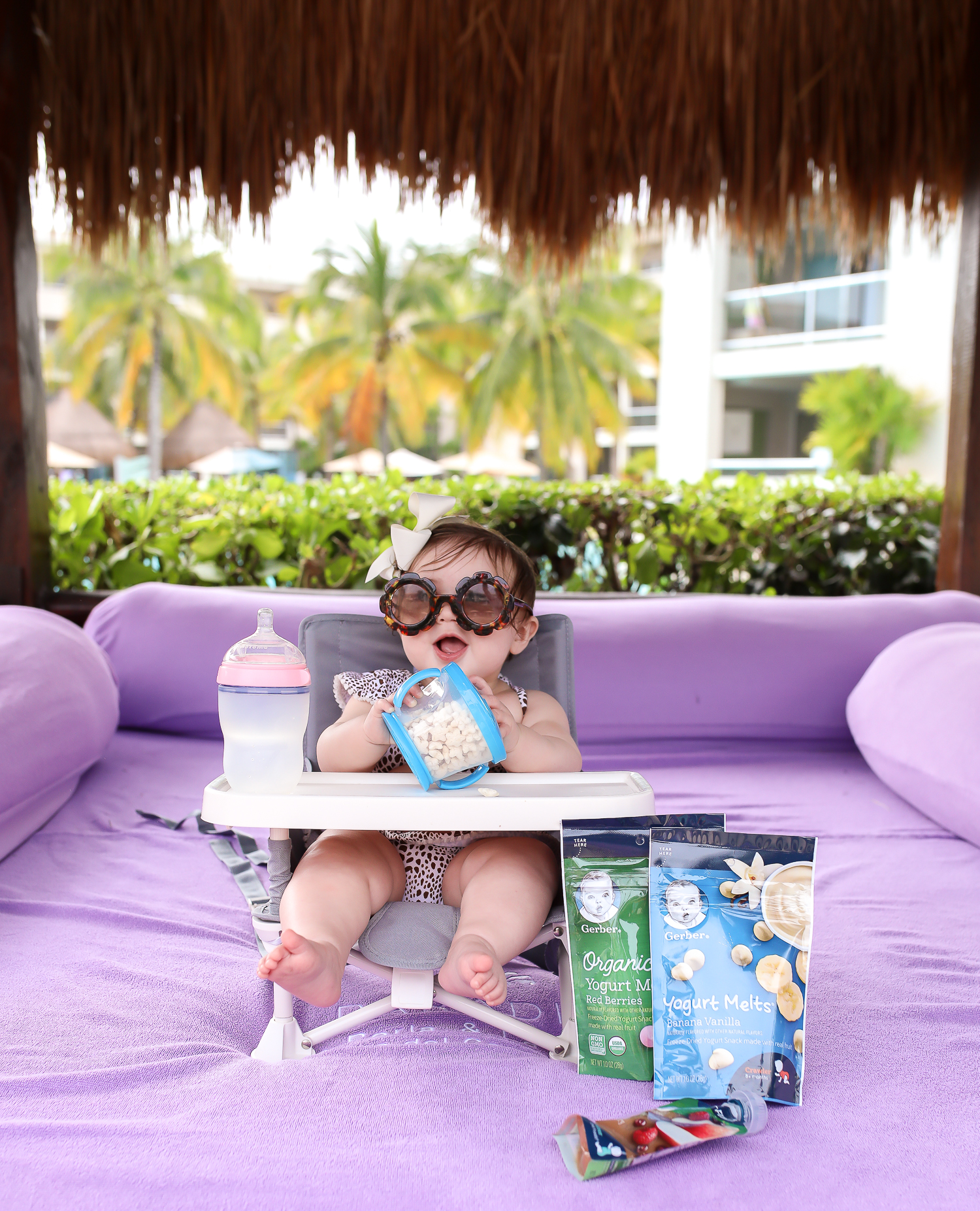 Christmas In Cancun🌟🎄🎁 [+ A Few Baby Travel Must-Haves] by popular Oklahoma travel blog, The Sweetest Thing: image of a baby in Cancun, Mexico sitting in a Walmart hiccapop Omniboost Travel Booster Seat with Tray for Baby holding a Walmart Munchkin Snack Catcher Snack Cup with Gerber Puff cereal snack inside, with a bottle resting on her high chair tray and some Gerber Organic Yogurt melts next to her high chair. 