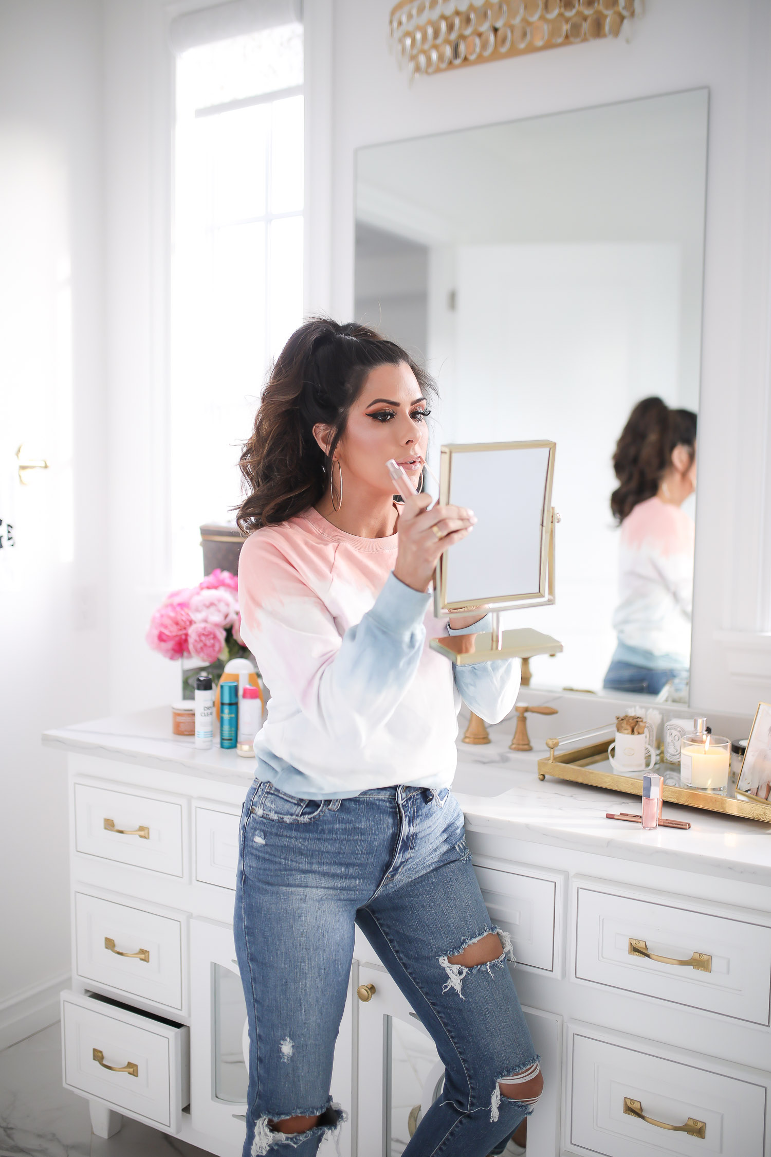 Sephora Favorites by popular US beauty blog, The Sweetest Thing: image of a woman wearing a tie dye sweatshirt and applying lip glass in a handheld mirror. 