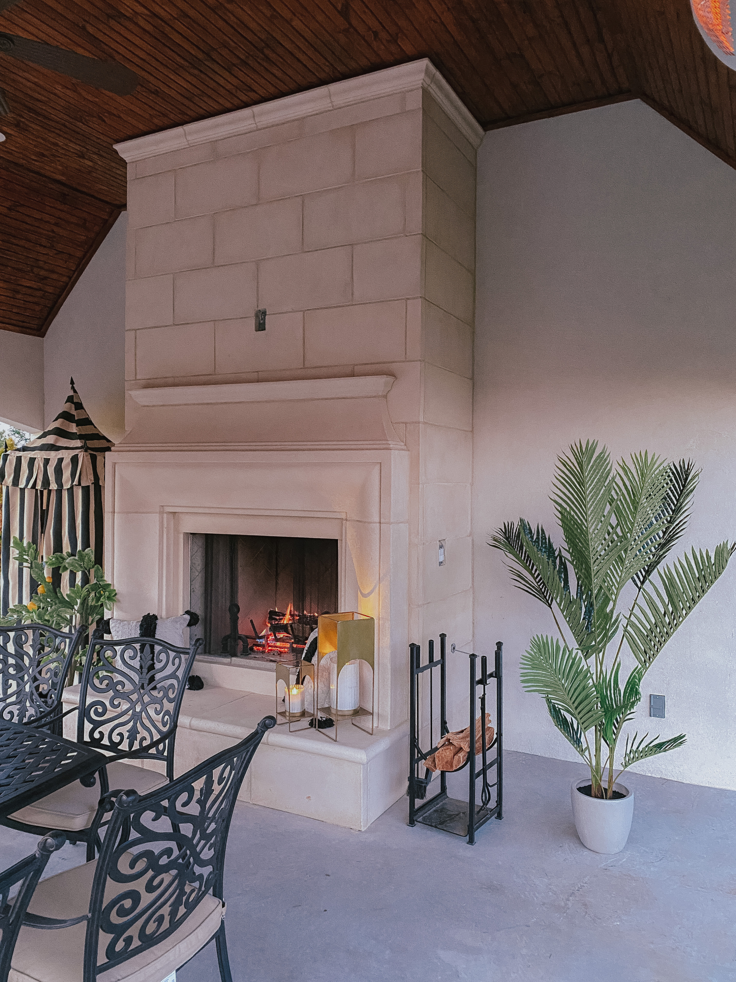 Backyard Decor by popular US life and style blog, The Sweetest Thing: image of a black metal outdoor table and chairs, faux palm tree plant, fireplace, metal log holder, and gold lanterns. 