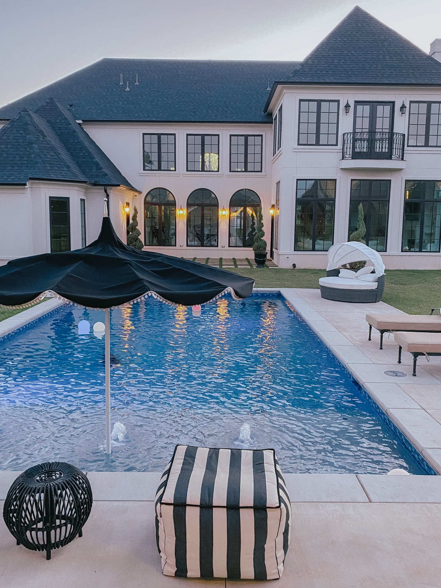 Backyard Decor by popular US life and style blog, The Sweetest Thing: image of a backyard pool, black and white stripe pouf, floating pool orbs, black umbrella with white pom-pom fringe, black and white strip lumbar pillows, and lounge chairs with tan cushions. 