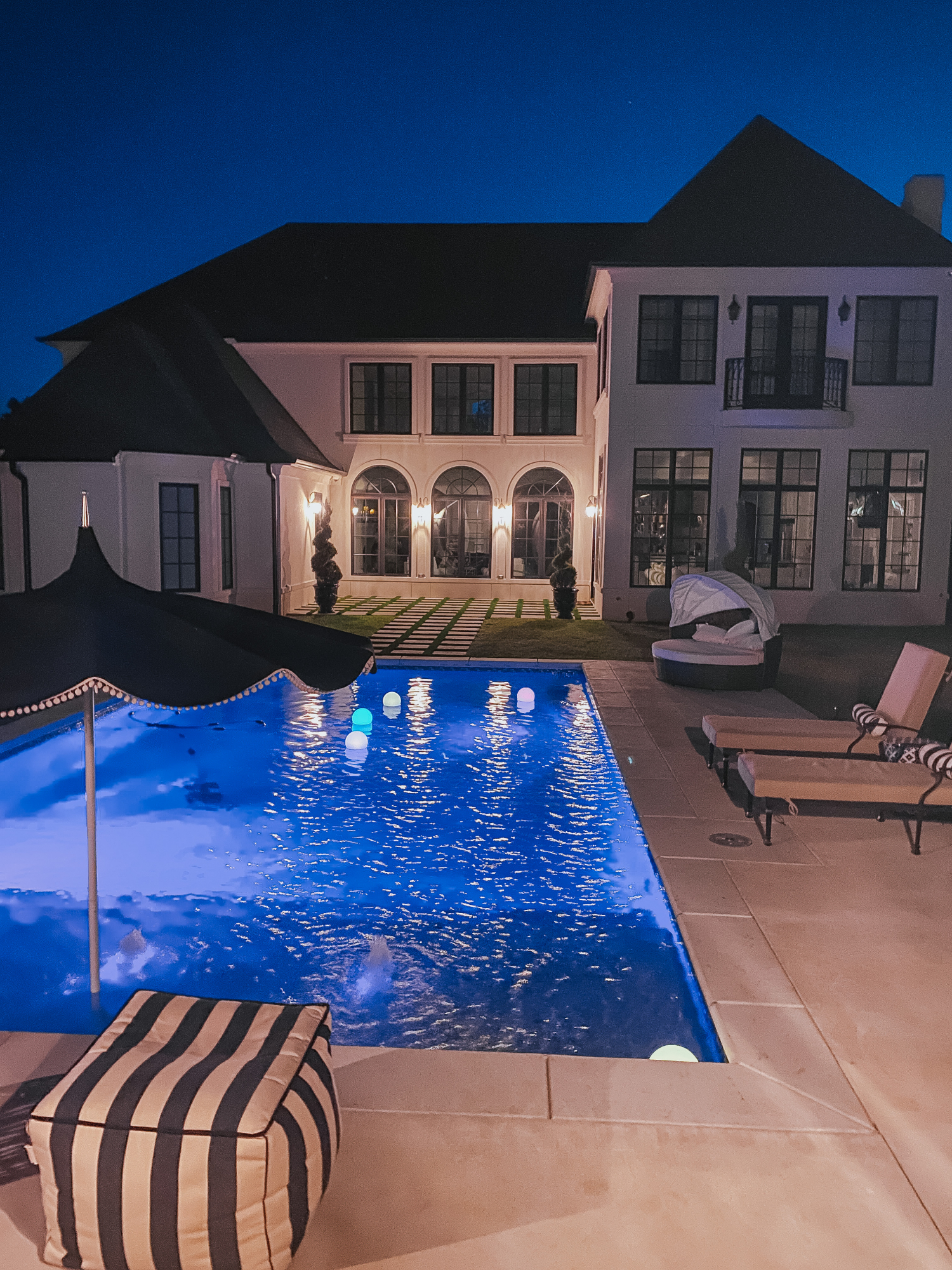 Backyard Decor by popular US life and style blog, The Sweetest Thing: image of a backyard pool, black and white stripe pouf, floating pool orbs, black umbrella with white pom-pom fringe, black and white strip lumbar pillows, and lounge chairs with tan cushions. 