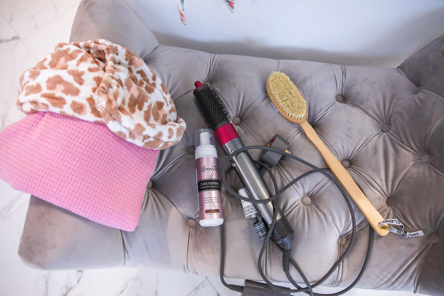 dyson airwrap review, master bathroom white and gold, Emily Gemma | Nighttime Routine by popular US beauty blog, The Sweetest Thing: image of a leopard print body towel, Elemis body exfoliating body brush, and pink textured towel on a grey velvet tuft bench. 