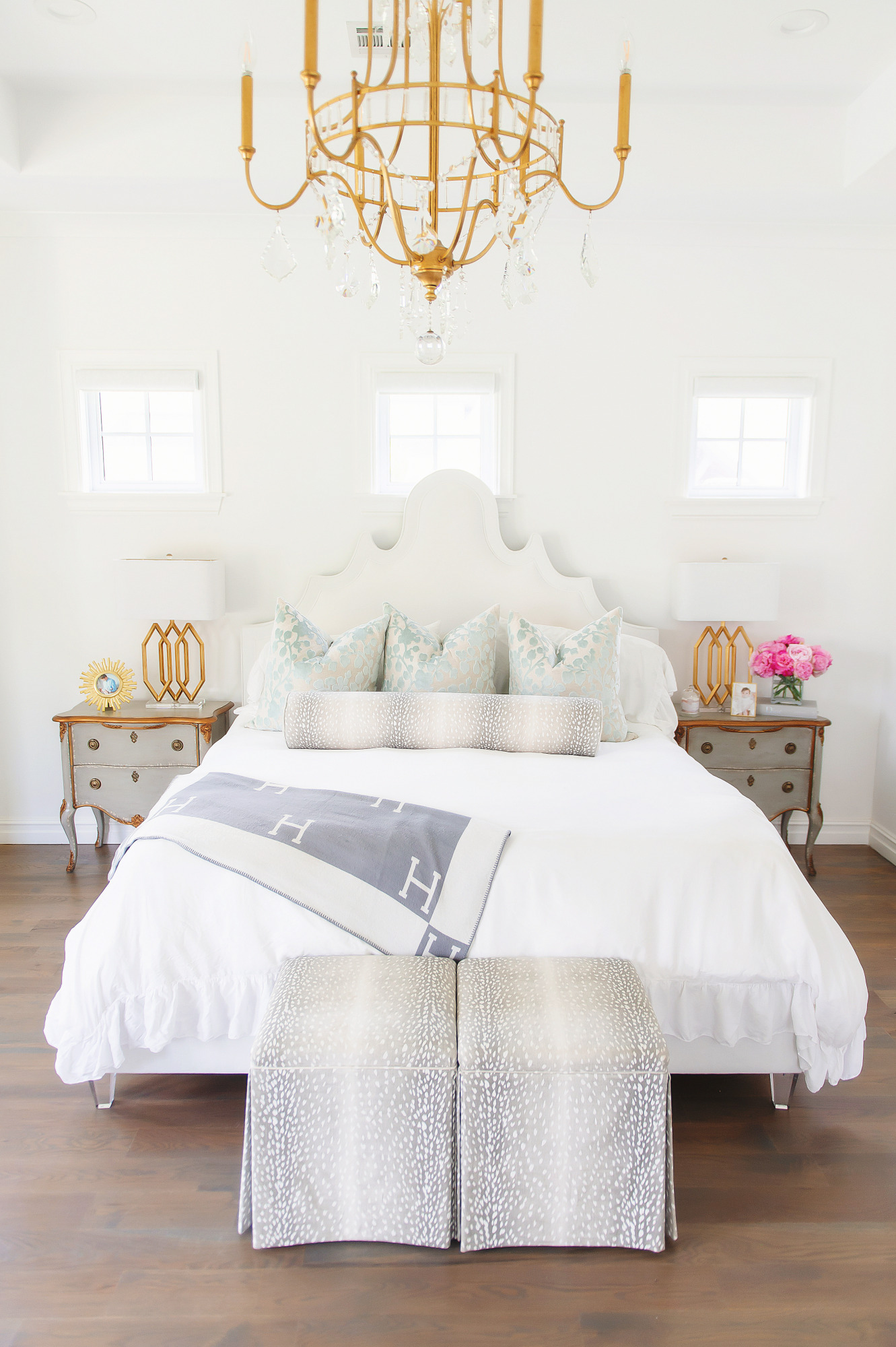 Blog Design by popular US lifestyle blog, The Sweetest Thing: image of Emily Gemma's master bedroom with a gold and crystal chandelier, grey wood night stands, antelope print bench, and a bed with a fabric headboard, antelope print lumbar pillow and white duvet cover. 