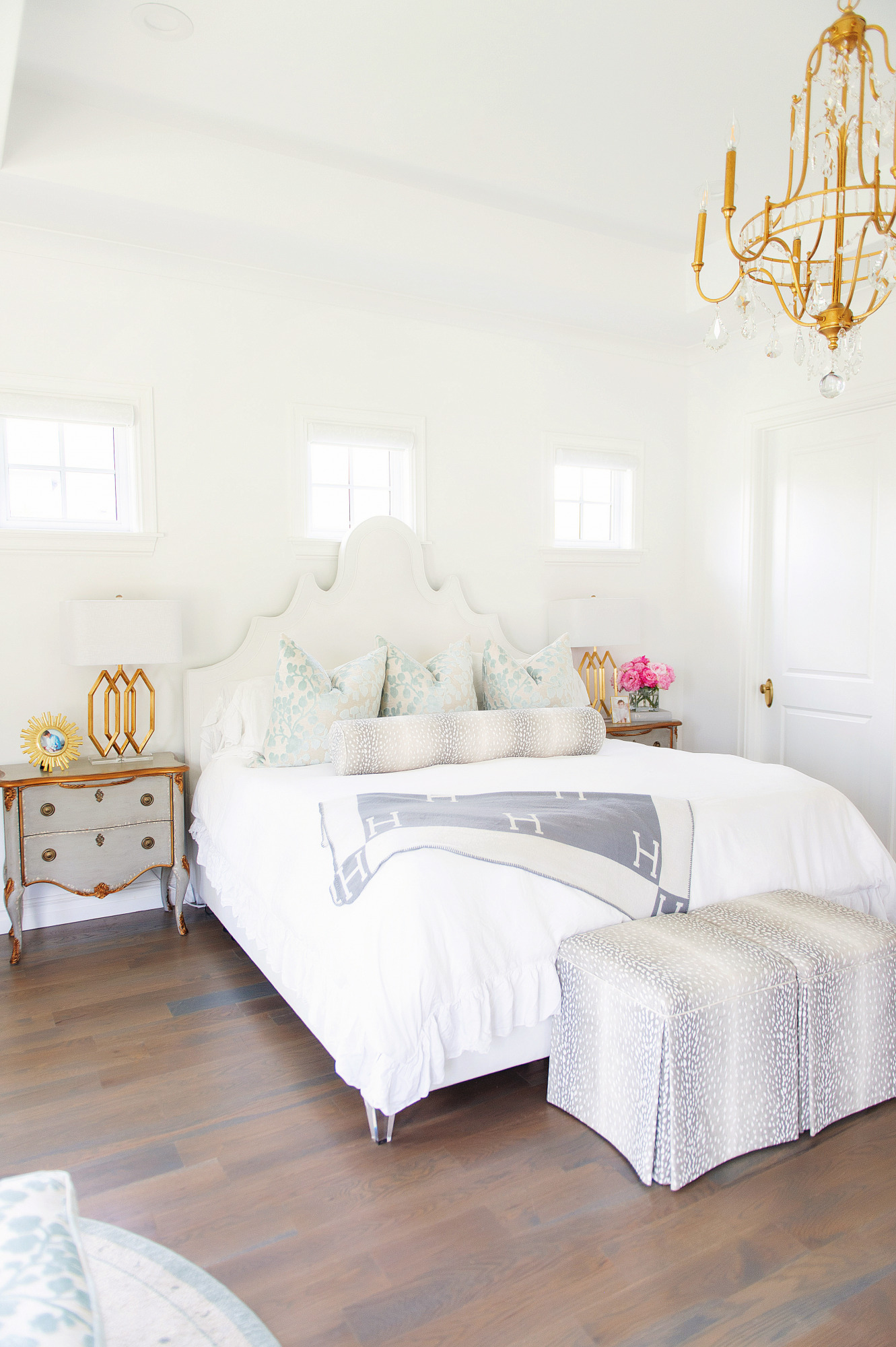 Blog Design by popular US lifestyle blog, The Sweetest Thing: image of Emily Gemma's master bedroom with a gold and crystal chandelier, grey wood night stands, antelope print bench, and a bed with a fabric headboard, antelope print lumbar pillow and white duvet cover. 