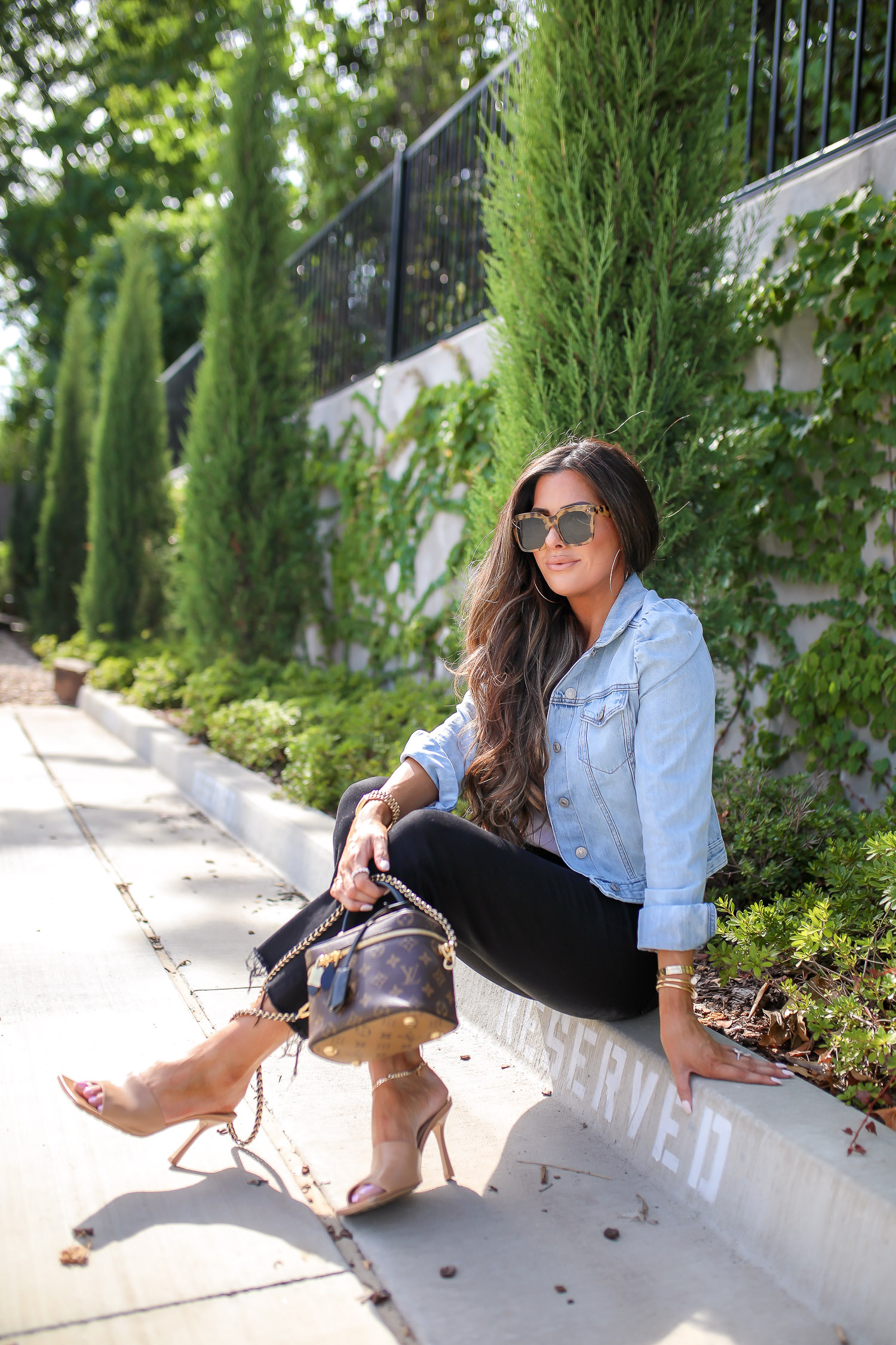 Denim Jacket by popular US fashion blog, The Sweetest Thing: image of Emily Gemma sitting outside in a parking lot and wearing a Express Puff Sleeve Light Wash Denim Jacket, Express Scoop Neck Sweater Cami, Express High Waisted Black Raw Hem Cropped Wide Leg Jeans, Express 2" Metal Hoop Earrings, Express Chain Ankle Bracelet, Cartier bracelets, Bottega Veneta shoes and holding a Louis Vuitton purse. 