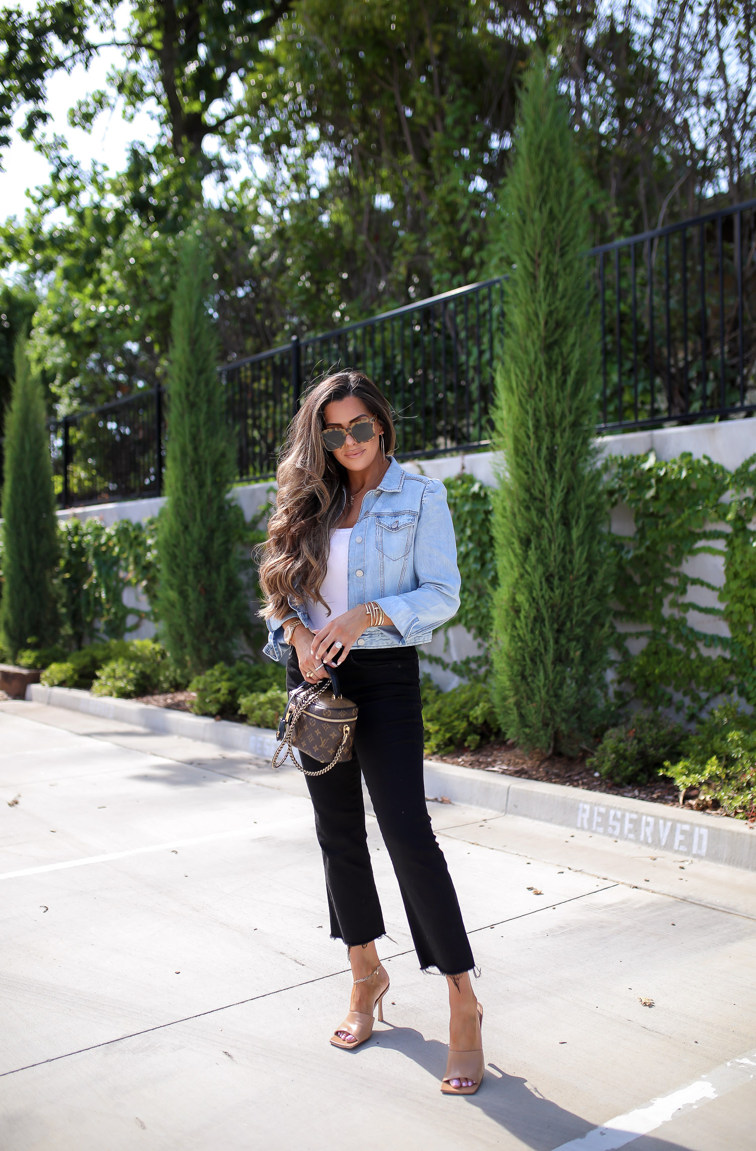 Denim Jacket by popular US fashion blog, The Sweetest Thing: image of Emily Gemma standing outside in a parking lot and wearing a Express Puff Sleeve Light Wash Denim Jacket, Express Scoop Neck Sweater Cami, Express High Waisted Black Raw Hem Cropped Wide Leg Jeans, Express 2" Metal Hoop Earrings, Express Chain Ankle Bracelet, Cartier bracelets, Bottega Veneta shoes and holding a Louis Vuitton purse. 