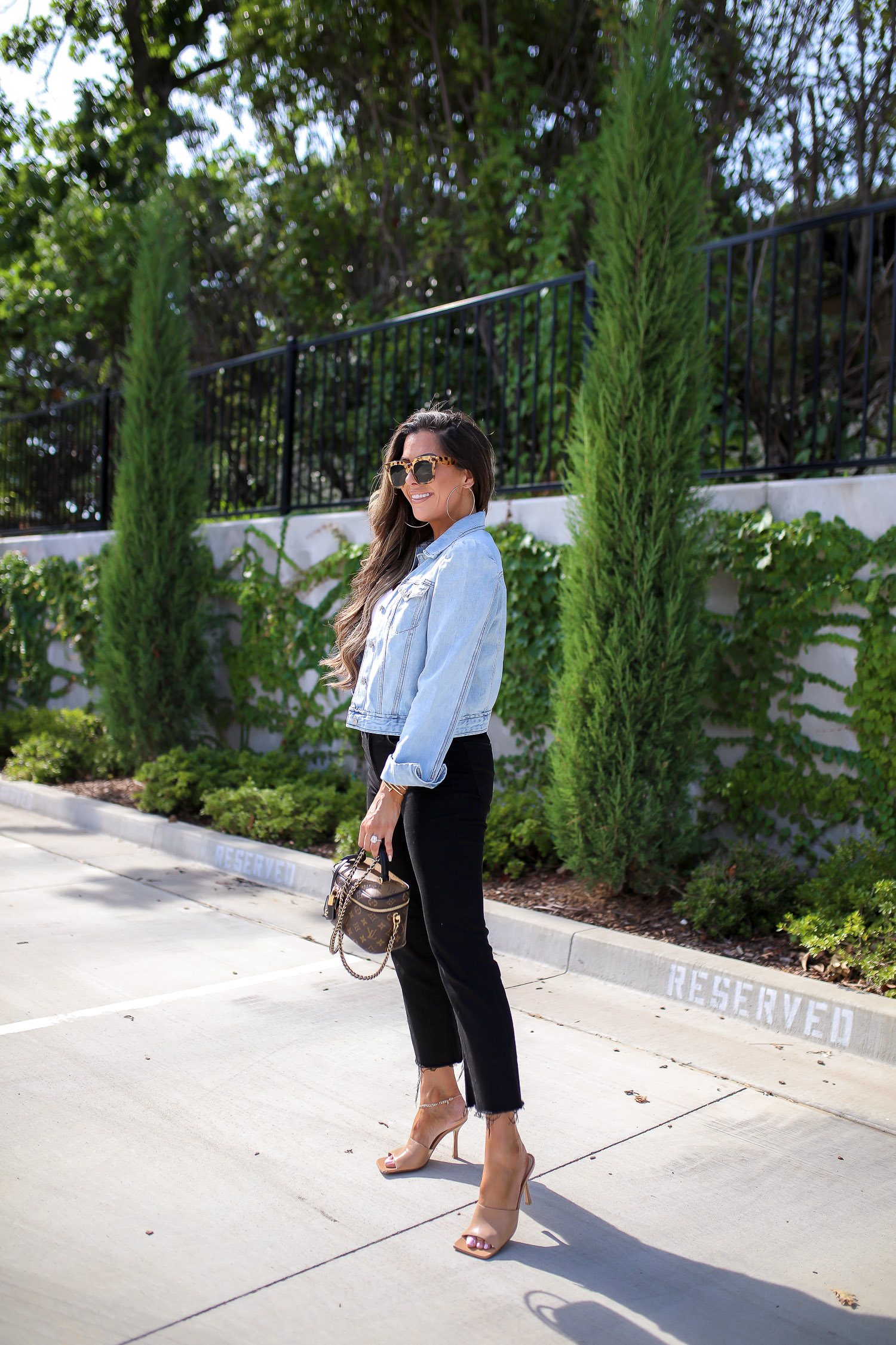 Denim Jacket by popular US fashion blog, The Sweetest Thing: image of Emily Gemma standing outside in a parking lot and wearing a Express Puff Sleeve Light Wash Denim Jacket, Express Scoop Neck Sweater Cami, Express High Waisted Black Raw Hem Cropped Wide Leg Jeans, Express 2" Metal Hoop Earrings, Express Chain Ankle Bracelet, Cartier bracelets, Bottega Veneta shoes and holding a Louis Vuitton purse. 