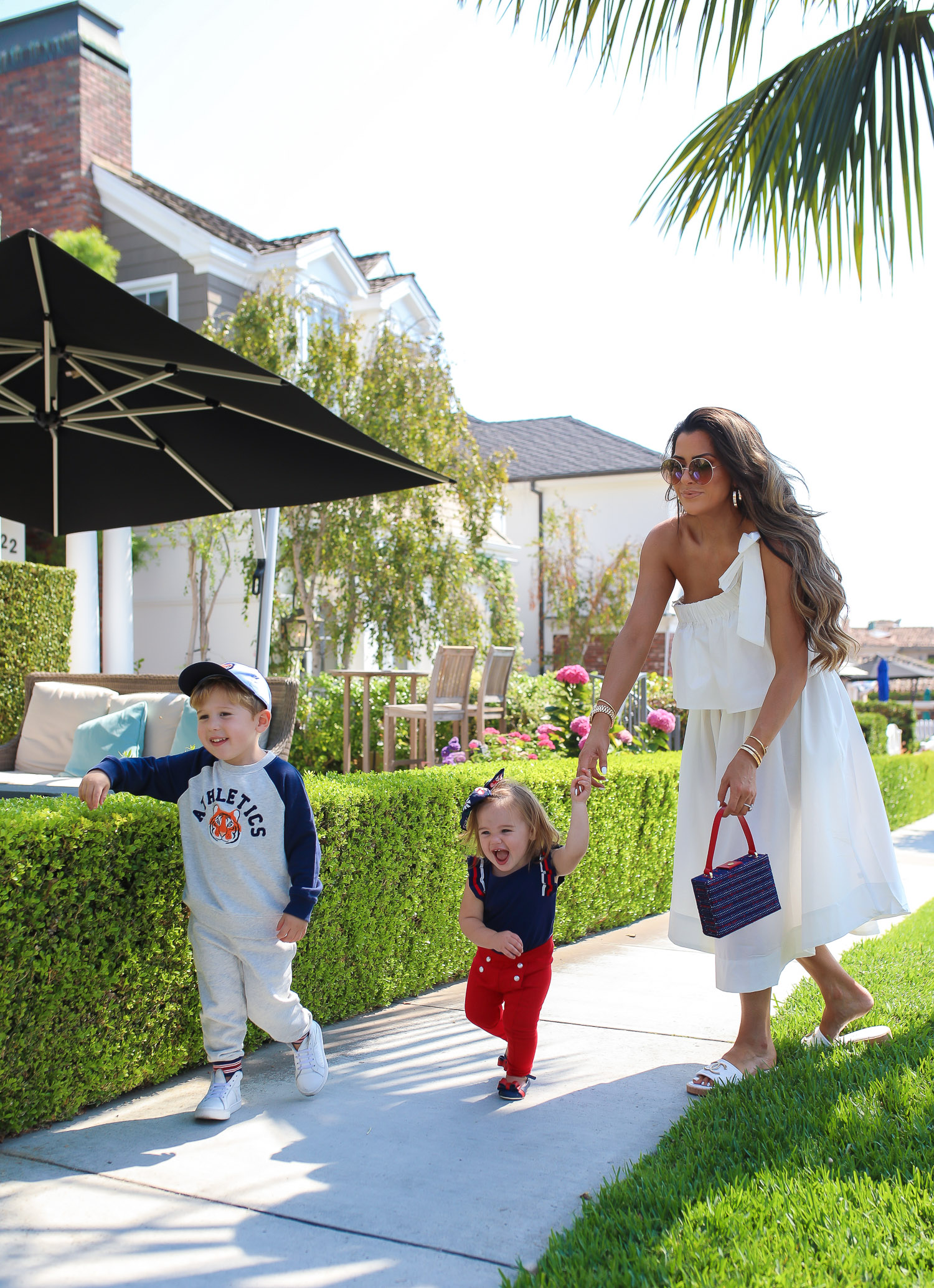 Janie and Jack by popular US fashion Blog, The Sweetest Thing: image of a mom and her two kids walking outside on a sidewalk in Newport Beach, CA and wearing a white one shoulder dress and white Channel slide sandals, Janie and Jack TIGER PATCH CAP, Janie and Jack RAGLAN TIGER SWEATSHIRT, Janie and Jack STRIPE TRIM JOGGER, Janie and Jack PLEATED SLEEVE TOP, Janie and Jack BUTTON PONTE PANT, Janie and Jack BOW ANKLE STRAP FLAT, FLORAL BOW BARRETTE, and Janie and Jack BOUCLÉ PURSE.