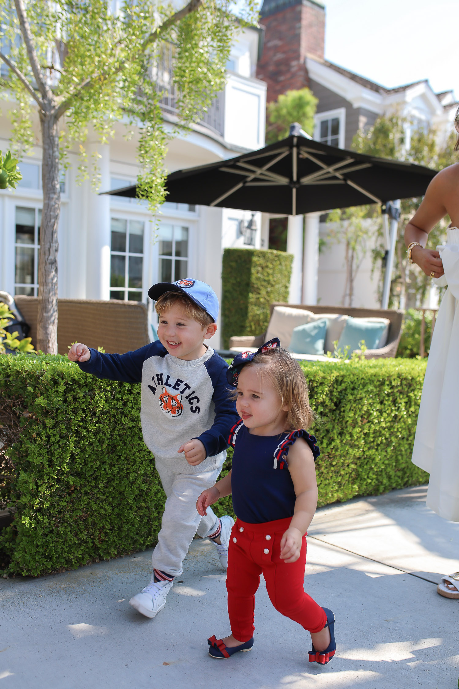 Janie and Jack by popular US fashion Blog, The Sweetest Thing: image of a mom and her two kids walking outside on a sidewalk in Newport Beach, CA and wearing a white one shoulder dress and white Channel slide sandals, Janie and Jack TIGER PATCH CAP, Janie and Jack RAGLAN TIGER SWEATSHIRT, Janie and Jack STRIPE TRIM JOGGER, Janie and Jack PLEATED SLEEVE TOP, Janie and Jack BUTTON PONTE PANT, Janie and Jack BOW ANKLE STRAP FLAT, FLORAL BOW BARRETTE, and Janie and Jack BOUCLÉ PURSE.