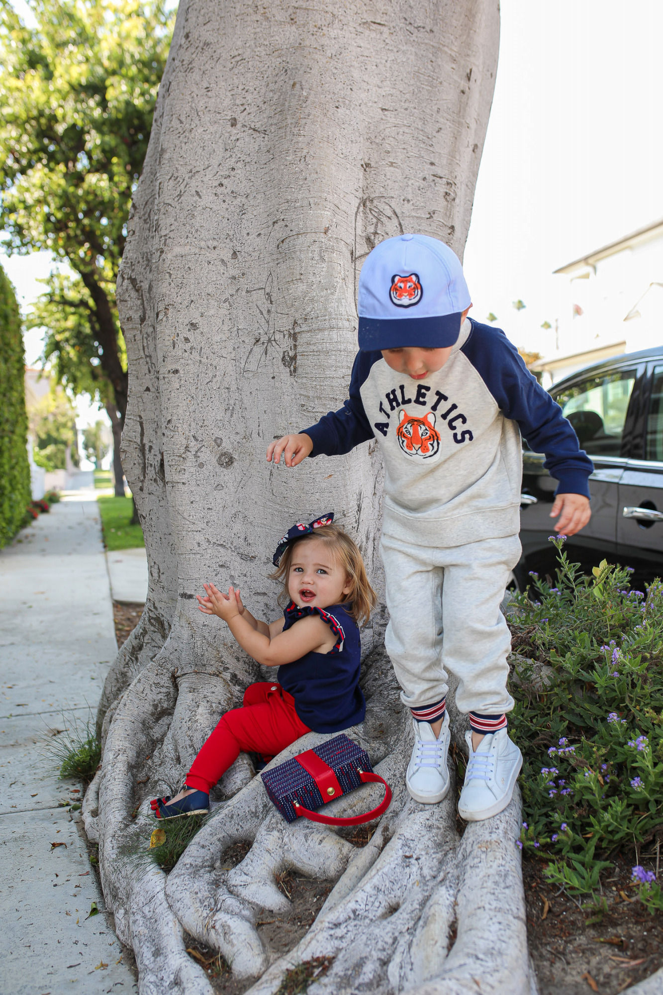 Janie and Jack by popular US fashion Blog, The Sweetest Thing: image of two kids walking outside on a sidewalk in Newport Beach, CA and wearing a Janie and Jack TIGER PATCH CAP, Janie and Jack RAGLAN TIGER SWEATSHIRT, Janie and Jack STRIPE TRIM JOGGER, Janie and Jack PLEATED SLEEVE TOP, Janie and Jack BUTTON PONTE PANT, Janie and Jack BOW ANKLE STRAP FLAT, FLORAL BOW BARRETTE, and Janie and Jack BOUCLÉ PURSE.