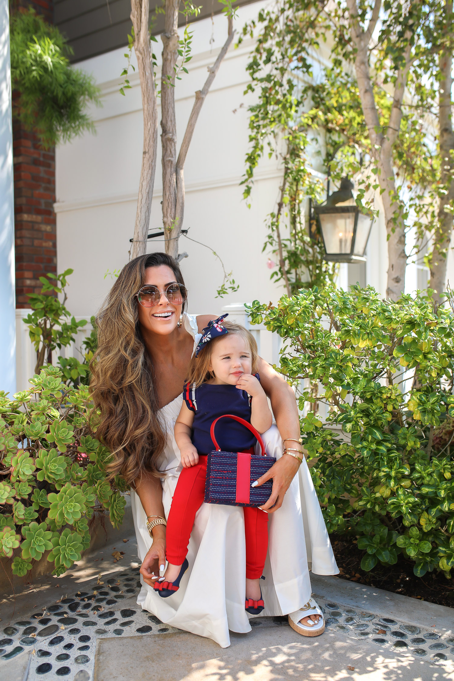 Janie and Jack by popular US fashion Blog, The Sweetest Thing: image of a mom holding her daughter outside in Newport Beach, CA and wearing a white one shoulder dress and white Channel slide sandals and Janie and Jack PLEATED SLEEVE TOP, Janie and Jack BUTTON PONTE PANT, Janie and Jack BOW ANKLE STRAP FLAT, FLORAL BOW BARRETTE, and Janie and Jack BOUCLÉ PURSE.