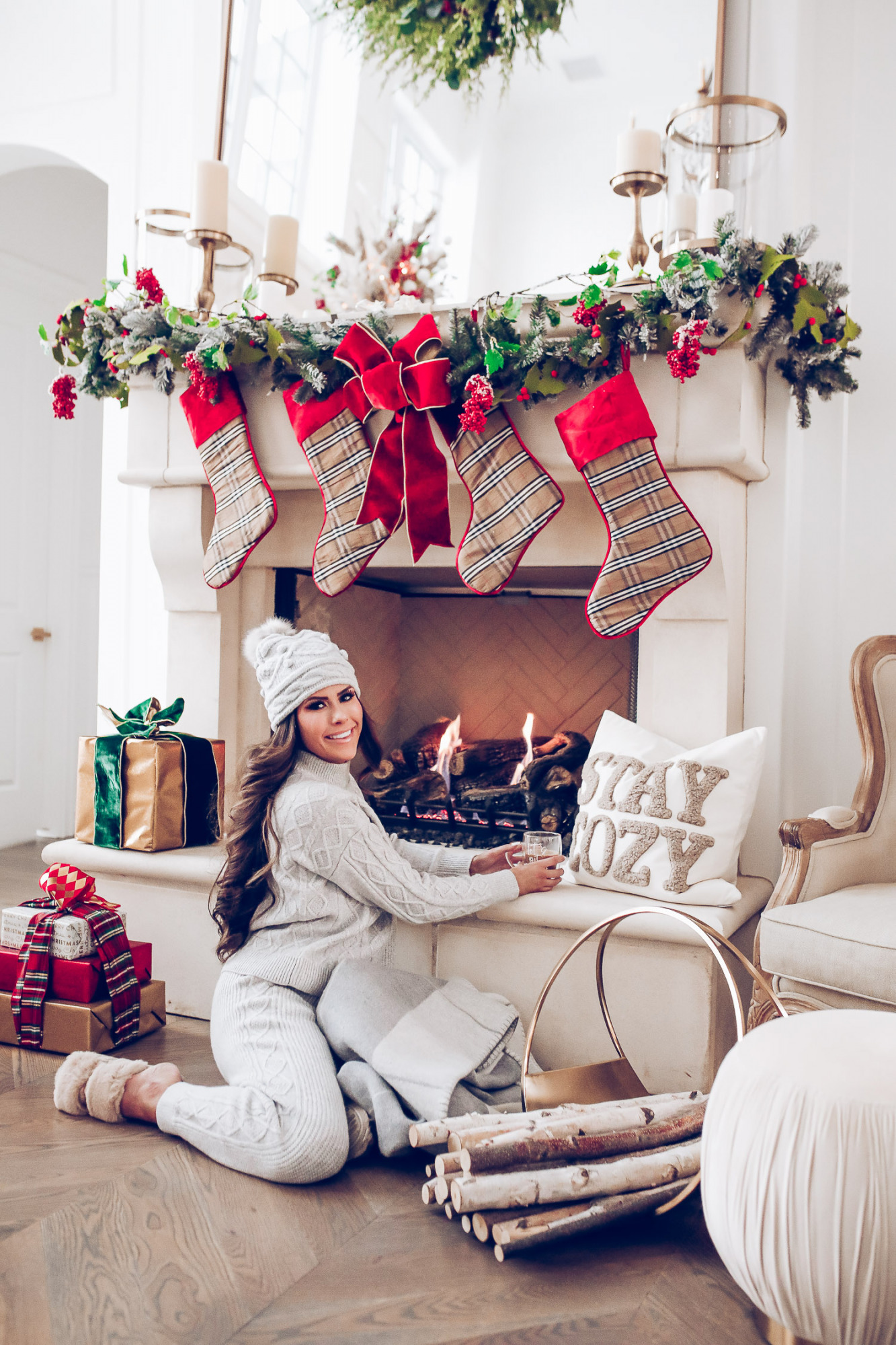 Express loungewear holiday 2020, express sweatsuit december 2020, emily gemma, holiday fireplace garland christmas |Cute Loungewear by popular US fashion blog, The Sweetest Thing: image of Emily Gemma sitting in front of a fireplace decorated with a garland, plaid stockings, and wrapped presents and wearing an Express sweater, Express pants, Express fuzzy slippers, and an Express pom beanie. 