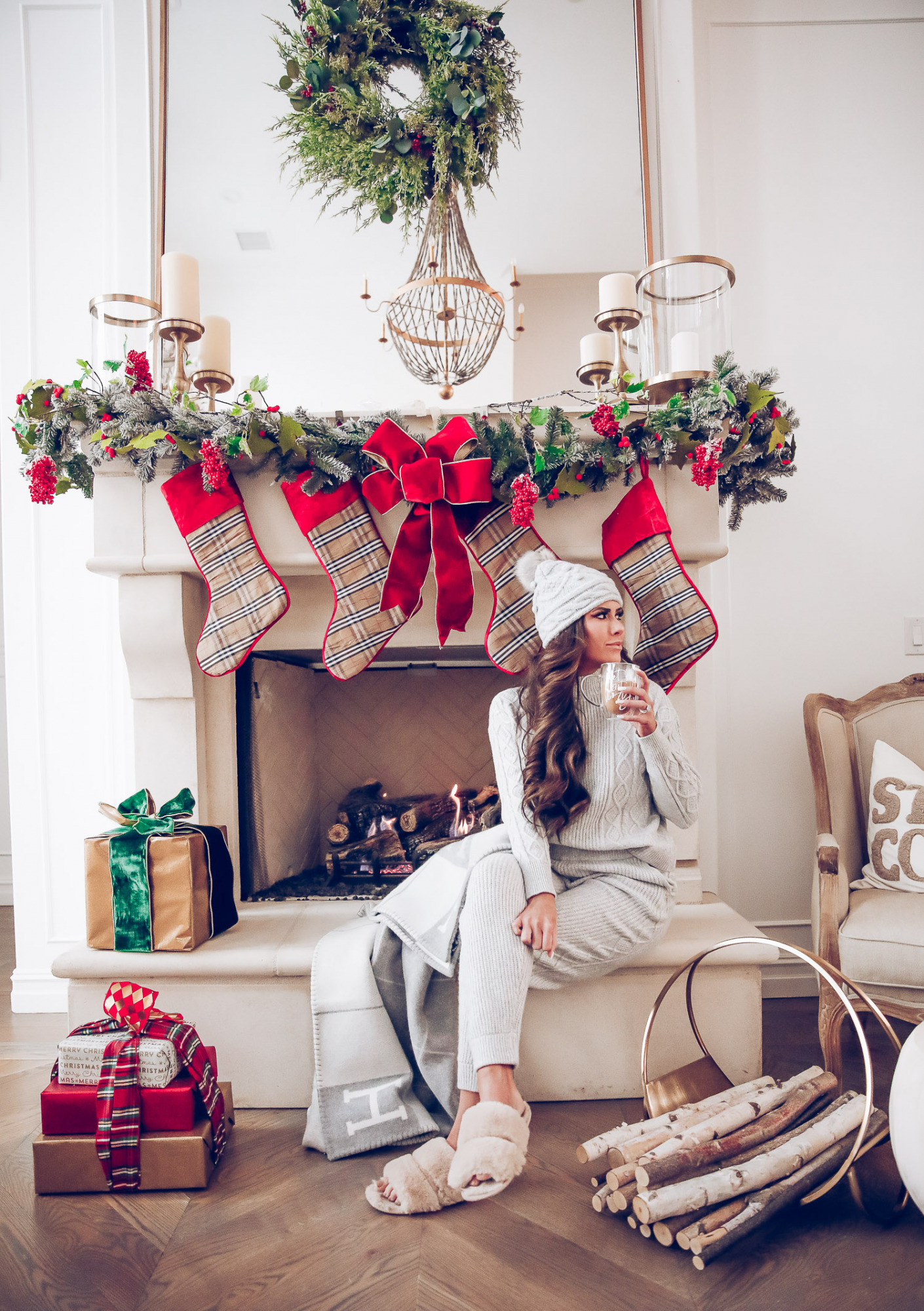 Express loungewear holiday 2020, express sweatsuit december 2020, emily gemma, holiday fireplace garland christmas | Cute Loungewear by popular US fashion blog, The Sweetest Thing: image of Emily Gemma sitting in front of a fireplace decorated with a garland, plaid stockings, and wrapped presents and wearing an Express sweater, Express pants, Express fuzzy slippers, and an Express pom beanie. 