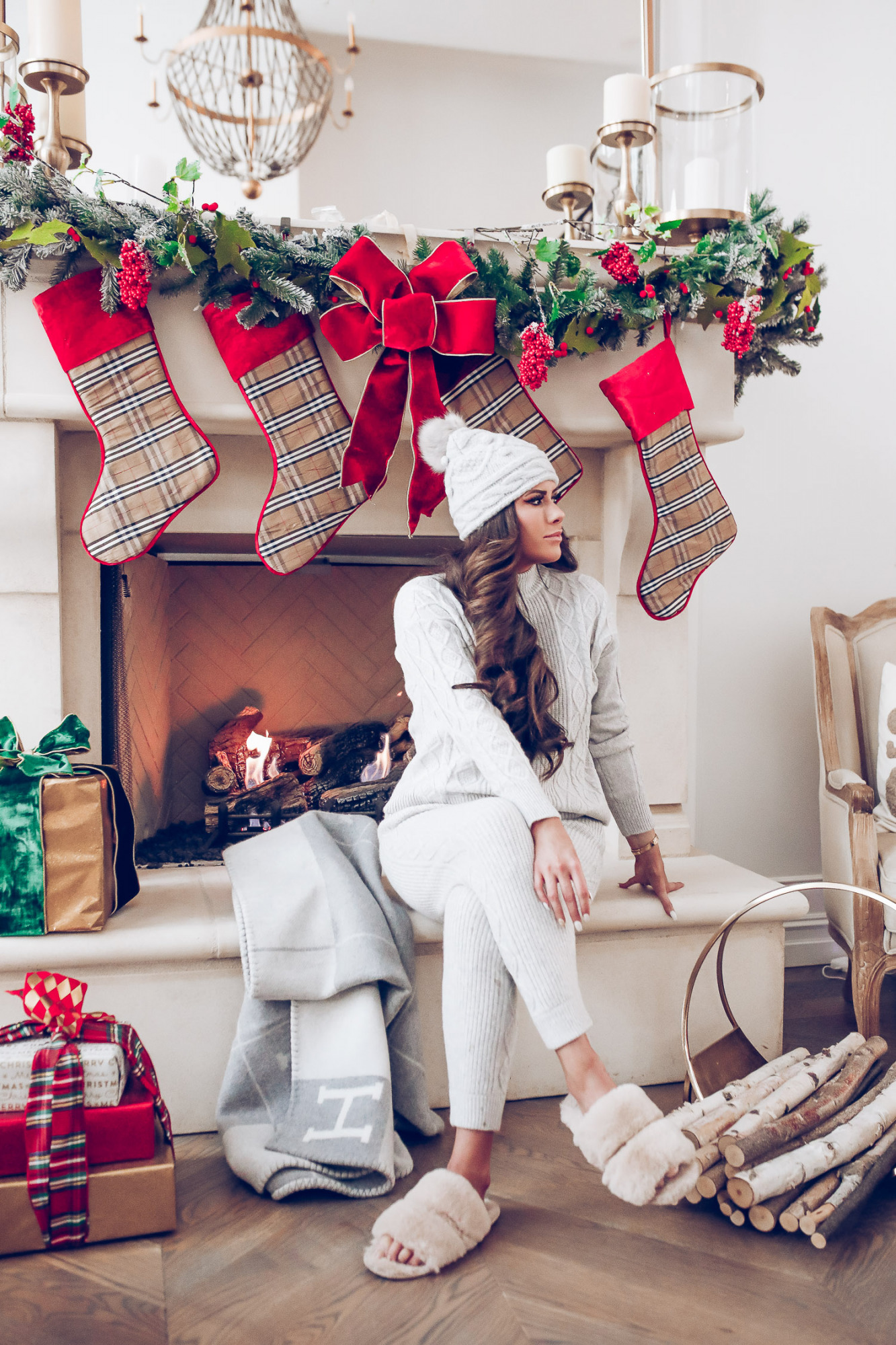 Express loungewear holiday 2020, express sweatsuit december 2020, emily gemma, holiday fireplace garland christmas |Cute Loungewear by popular US fashion blog, The Sweetest Thing: image of Emily Gemma sitting in front of a fireplace decorated with a garland, plaid stockings, and wrapped presents and wearing an Express sweater, Express pants, Express fuzzy slippers, and an Express pom beanie. 