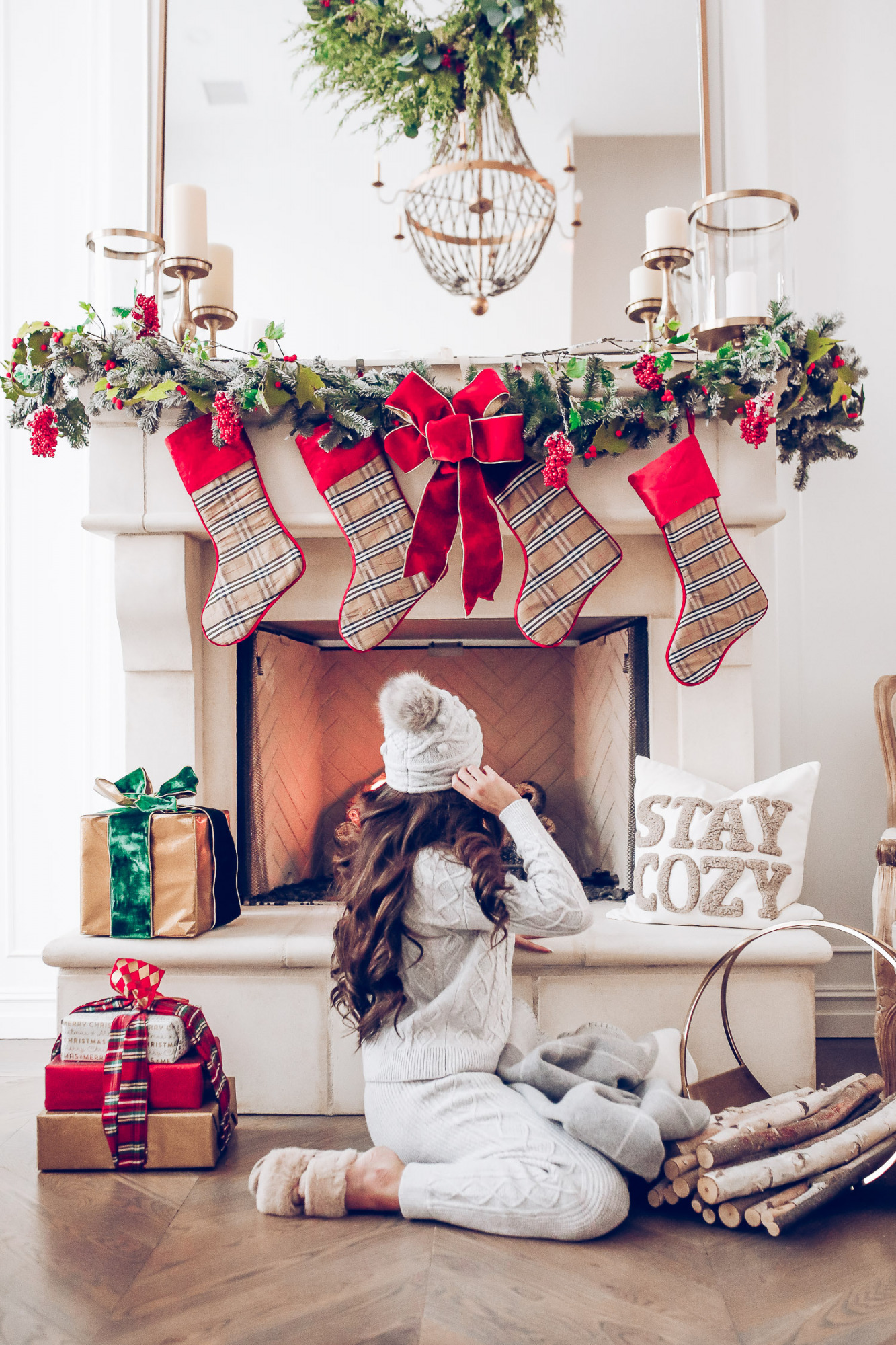 Express loungewear holiday 2020, express sweatsuit december 2020, emily gemma, holiday fireplace garland christmas |Cute Loungewear by popular US fashion blog, The Sweetest Thing: image of Emily Gemma sitting in front of a fireplace decorated with a garland, plaid stockings, and wrapped presents and wearing an Express sweater, Express pants, Express fuzzy slippers, and an Express pom beanie.