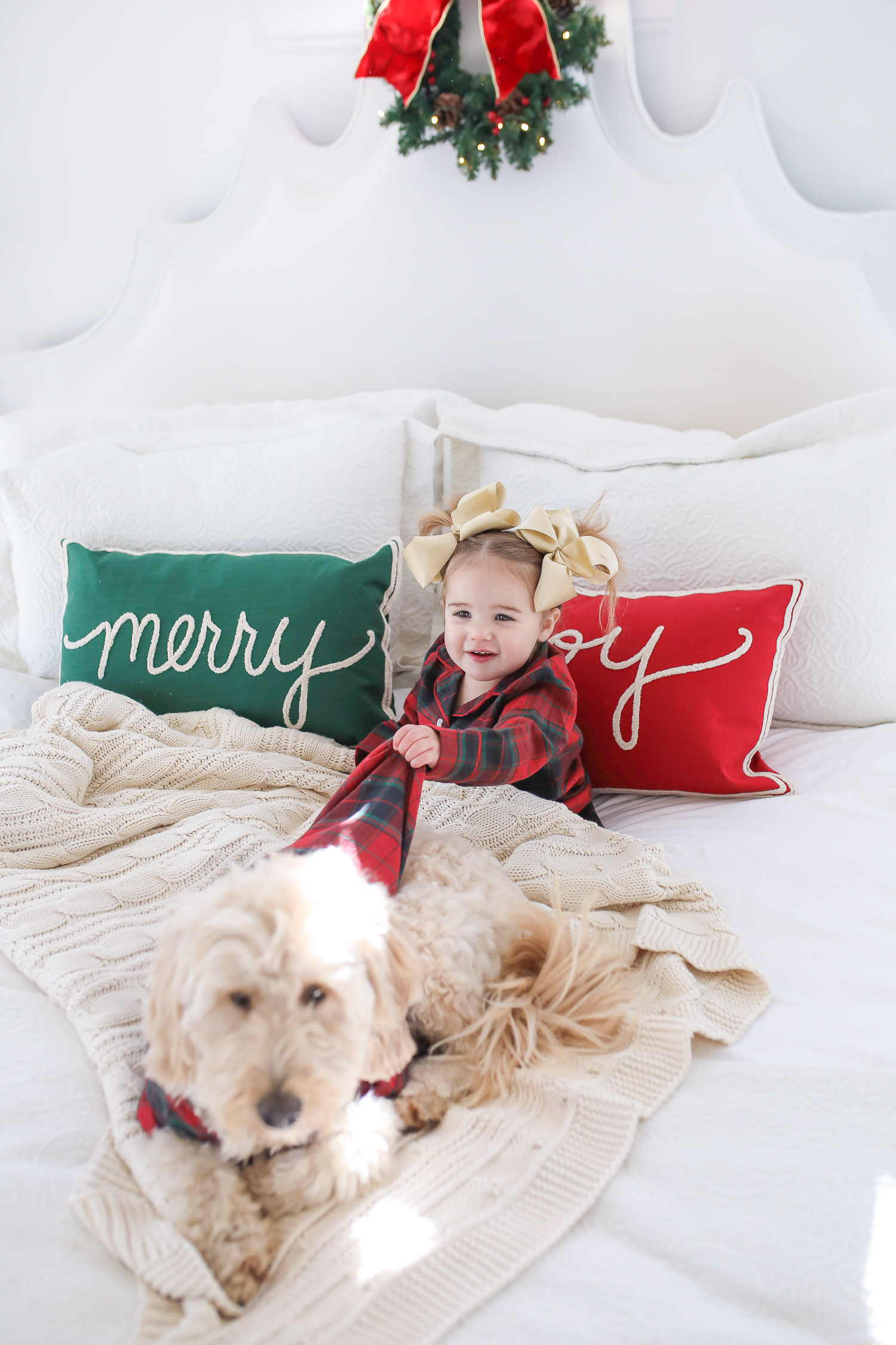 Matching Family Pajamas by popular US fashion blog, The Sweetest Thing: image of a young girl and her dog sitting on a bed with white bedding, a joy throw pillow, a merry throw pillow and a mini Christmas wreath hanging on the headboard and wearing a The Company Store Family Flannel Company Cotton™ Girls’ Sleepshirt and The Company Store Family Flannel Company Cotton™ Dog Pajamas.