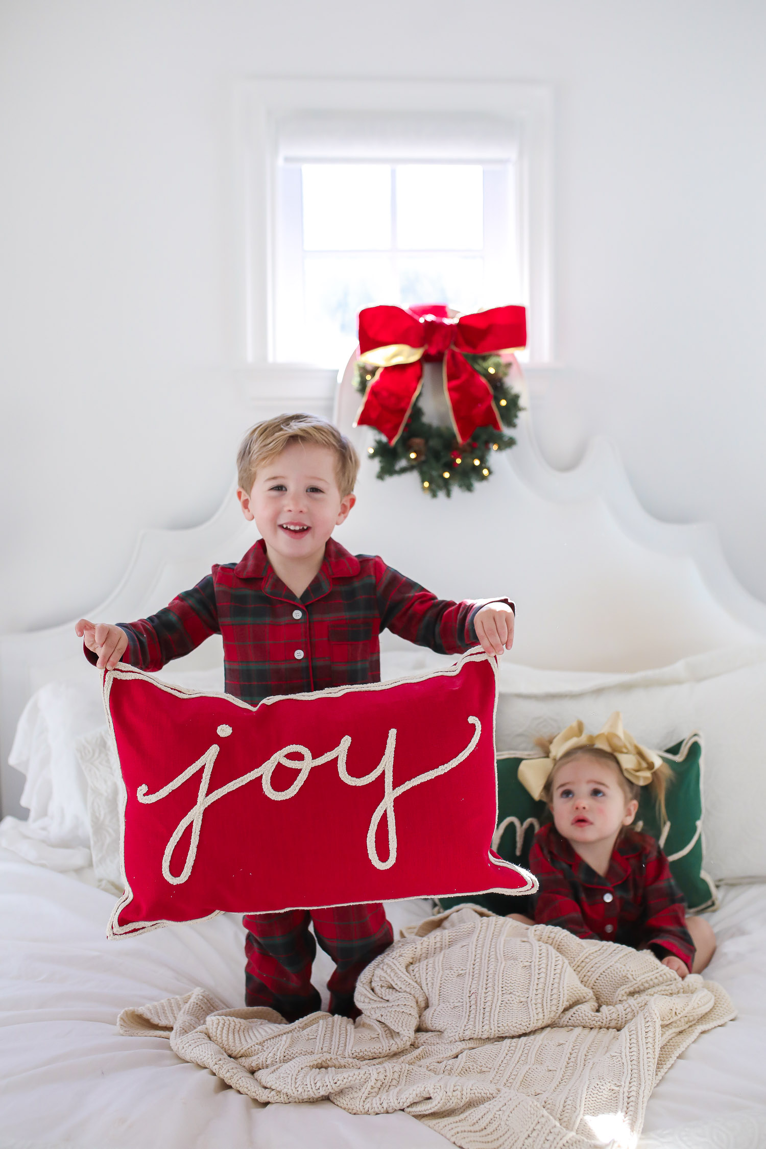 Matching Family Pajamas by popular US fashion blog, The Sweetest Thing: image of a young boy and girl sitting on a bed with white bedding, a joy throw pillow, a merry throw pillow and a mini Christmas wreath hanging on the headboard and wearing a The Company Store Family Flannel Company Cotton™ Girls’ Sleepshirt, The Company Store Family Flannel Company Cotton™ Kids’ Pajama Set.