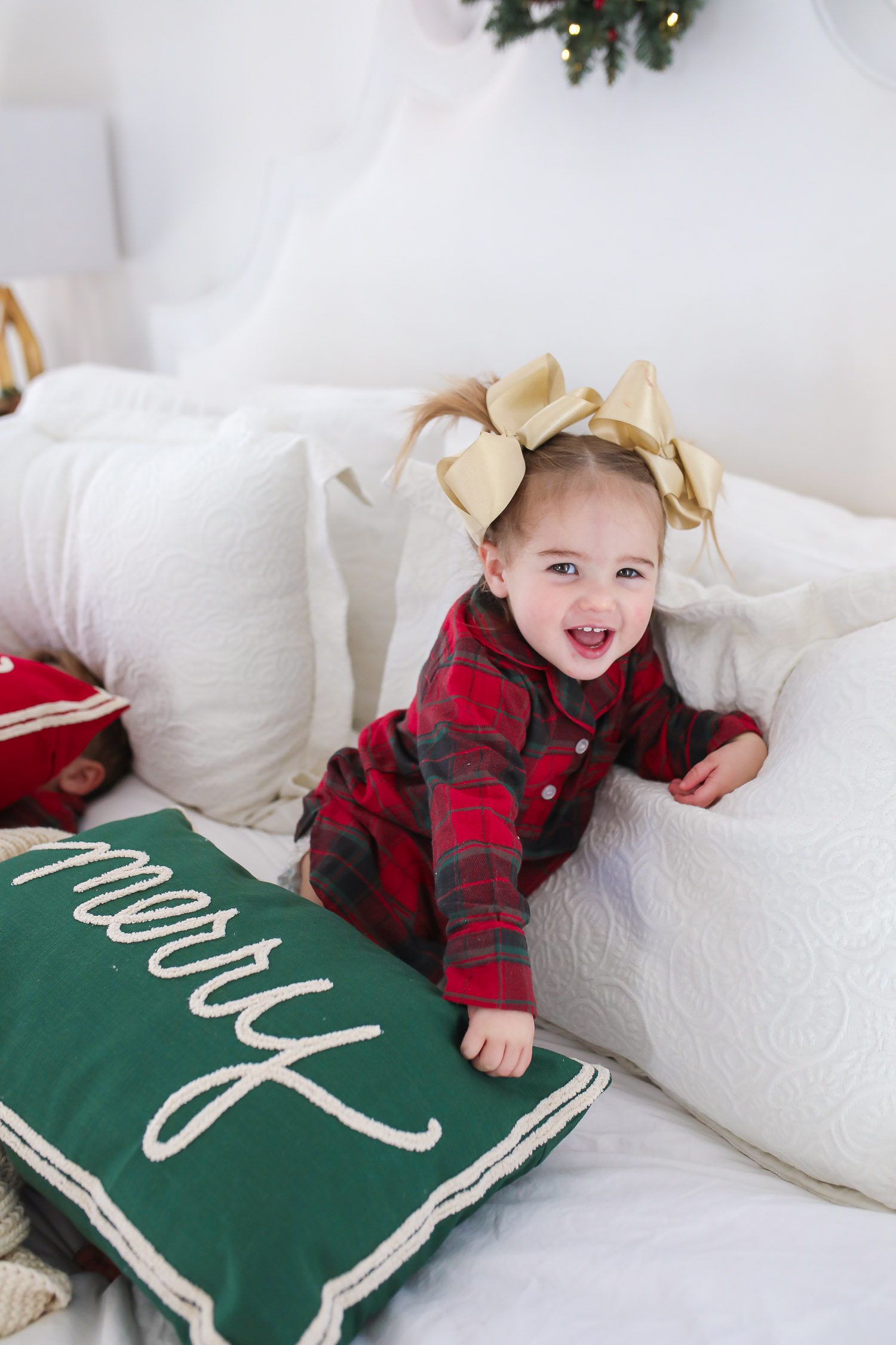 Matching Family Pajamas by popular US fashion blog, The Sweetest Thing: image of a young girl sitting on a bed with white bedding, a joy throw pillow, a merry throw pillow and a mini Christmas wreath hanging on the headboard and wearing a The Company Store Family Flannel Company Cotton™ Girls’ Sleepshirt.