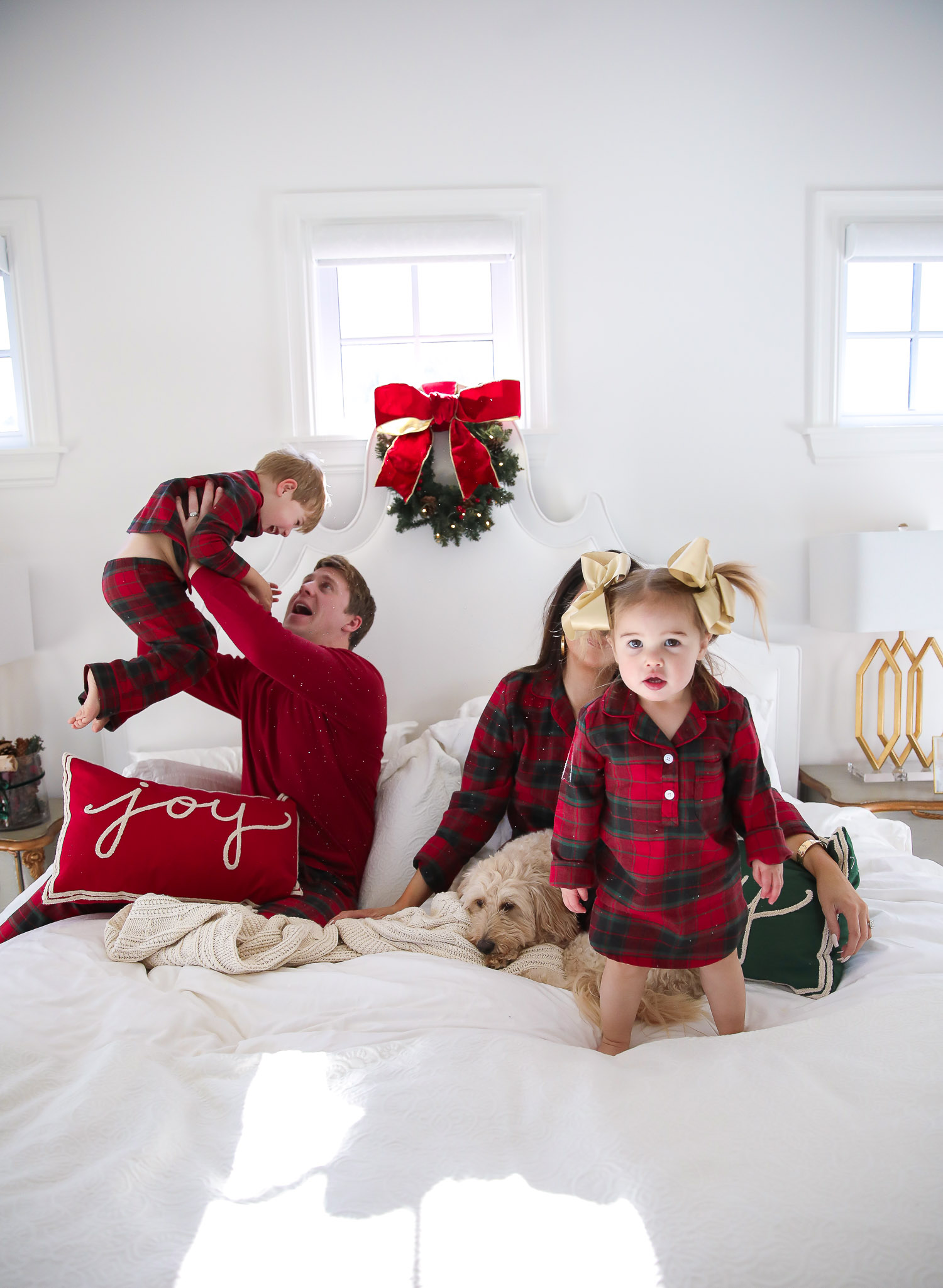 Matching Family Pajamas by popular US fashion blog, The Sweetest Thing: image of a family sitting on a bed with white bedding, a joy throw pillow, a merry throw pillow and a mini Christmas wreath hanging on the headboard and wearing a The Company Store Family Flannel Company Cotton™ Womens Pajama Set, The Company Store Family Flannel Company Cotton™ Girls’ Sleepshirt, The Company Store Family Flannel Company Cotton™ Kids’ Pajama Set, The Company Store Family Flannel Company Cotton™ Mens Pajama Set, and The Company Store Family Flannel Company Cotton™ Dog Pajamas.