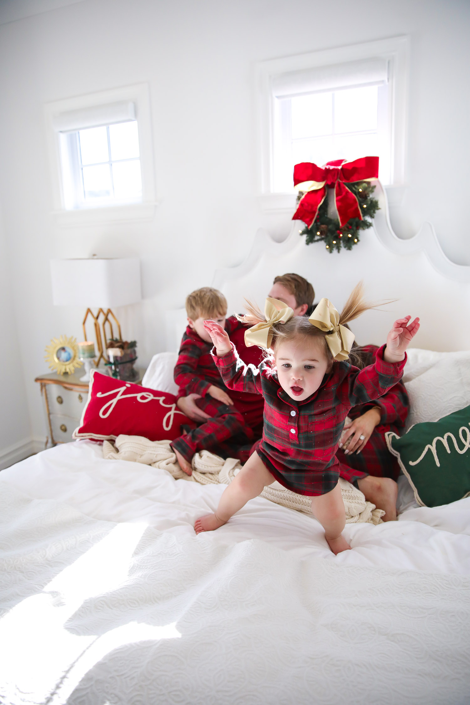Matching Family Pajamas by popular US fashion blog, The Sweetest Thing: image of a family sitting on a bed with white bedding, a joy throw pillow, a merry throw pillow and a mini Christmas wreath hanging on the headboard and wearing a The Company Store Family Flannel Company Cotton™ Womens Pajama Set, The Company Store Family Flannel Company Cotton™ Girls’ Sleepshirt, The Company Store Family Flannel Company Cotton™ Kids’ Pajama Set, The Company Store Family Flannel Company Cotton™ Mens Pajama Set, and The Company Store Family Flannel Company Cotton™ Dog Pajamas.