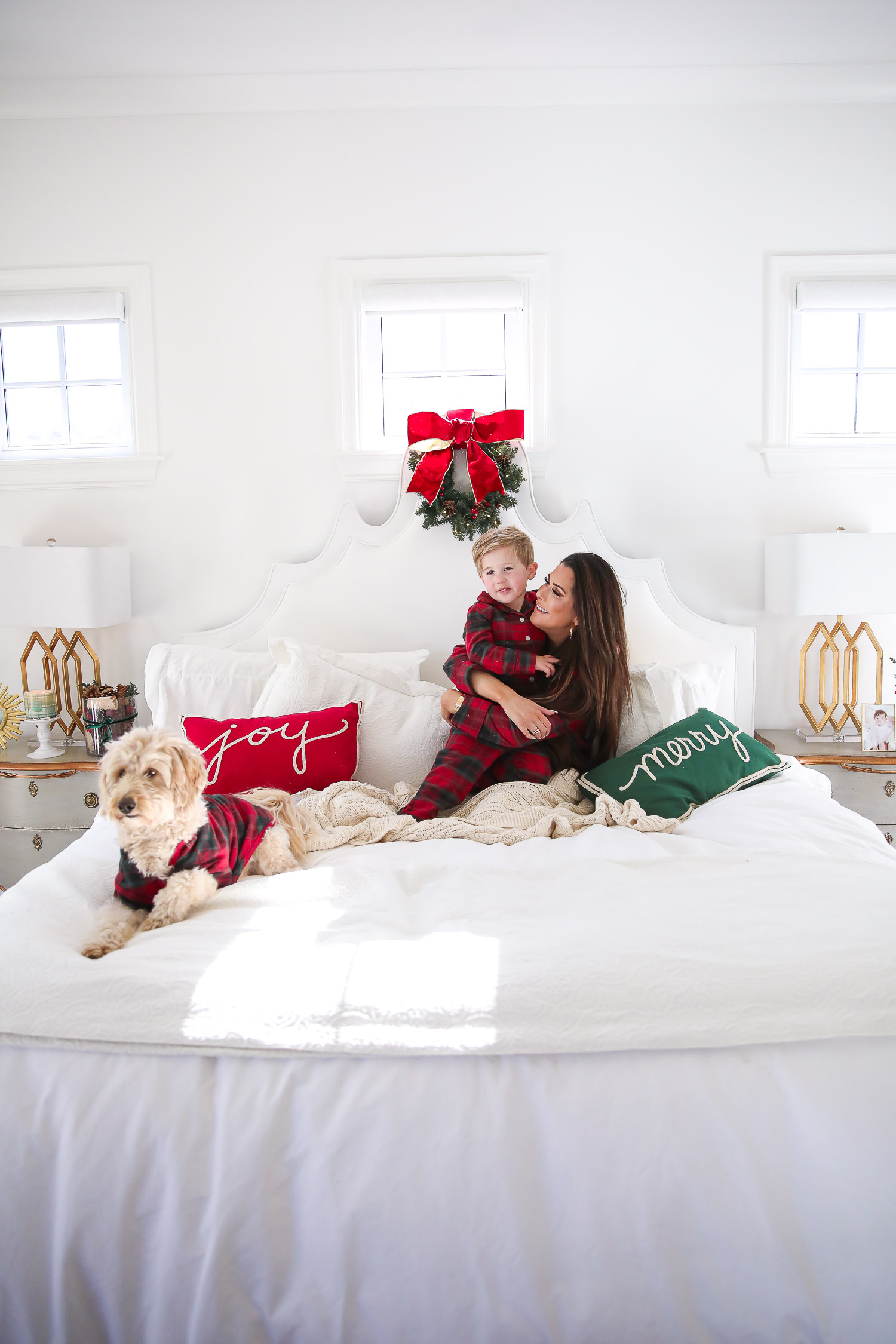 Matching Family Pajamas by popular US fashion blog, The Sweetest Thing: image of a Emily Gemma and her son sitting on a bed with white bedding, a joy throw pillow, a merry throw pillow and a mini Christmas wreath hanging on the headboard with their dog and wearing a The Company Store Family Flannel Company Cotton™ Womens Pajama Set, The Company Store Family Flannel Company Cotton™ Kids’ Pajama Set, and The Company Store Family Flannel Company Cotton™ Dog Pajamas.