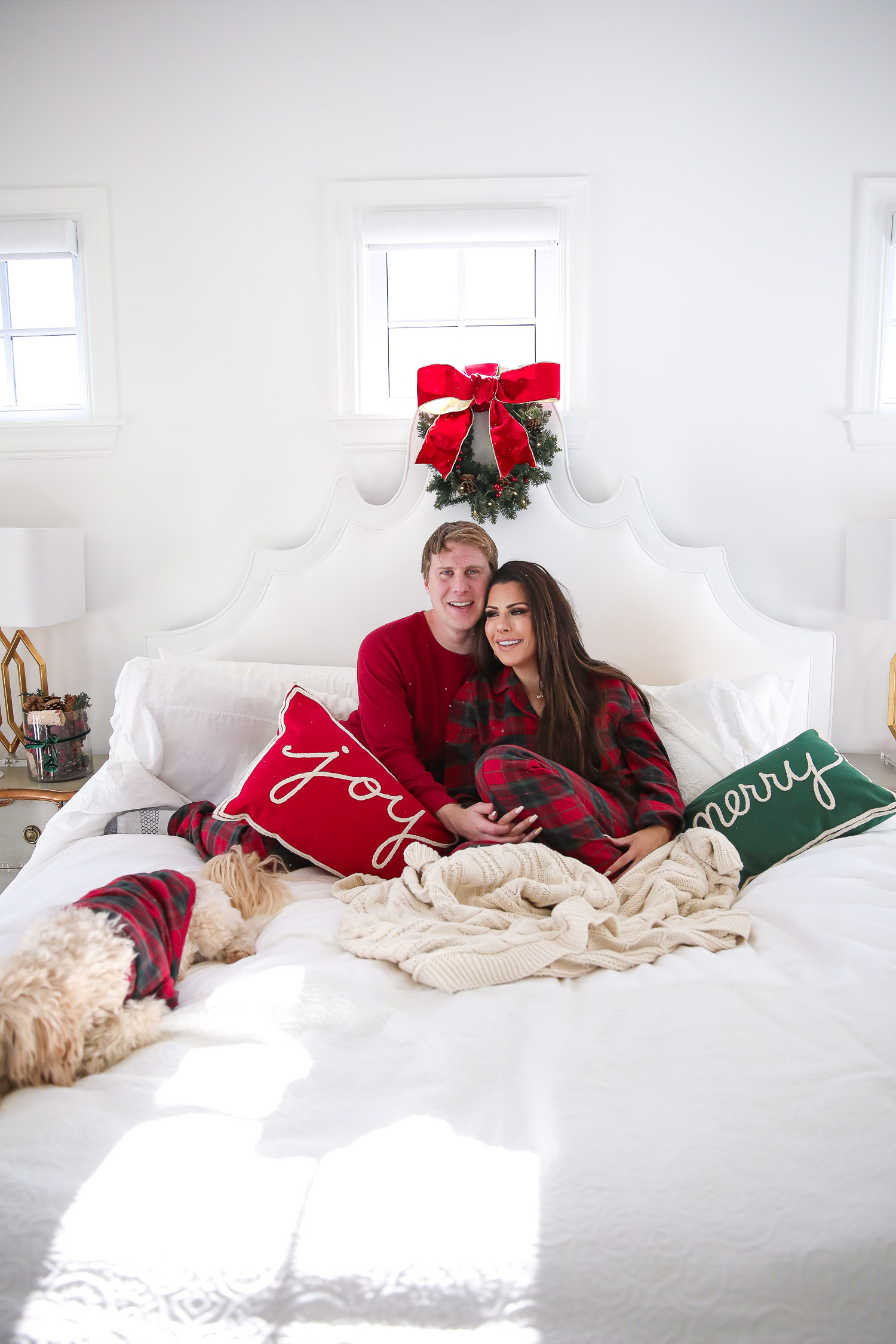 Matching Family Pajamas by popular US fashion blog, The Sweetest Thing: image of Emily Gemma and her husband John sitting on a bed with white bedding, a joy throw pillow, a merry throw pillow and a mini Christmas wreath hanging on the headboard with their dog and wearing a The Company Store Family Flannel Company Cotton™ Womens Pajama Set, The Company Store Family Flannel Company Cotton™ Mens Pajama Set, and The Company Store Family Flannel Company Cotton™ Dog Pajamas.