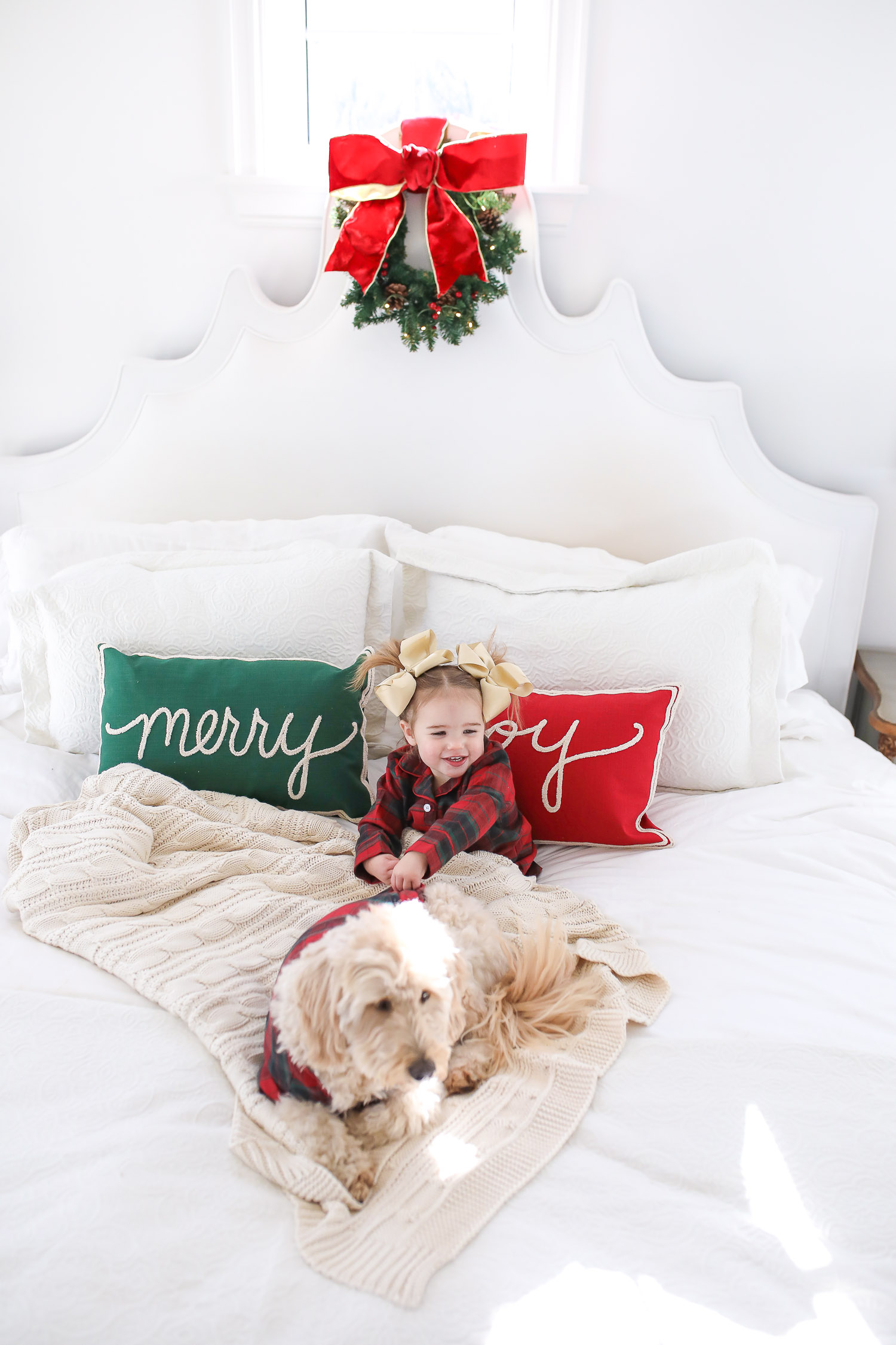 Matching Family Pajamas by popular US fashion blog, The Sweetest Thing: image of a young girl and her dog sitting on a bed with white bedding, a joy throw pillow, a merry throw pillow and a mini Christmas wreath hanging on the headboard and wearing a The Company Store Family Flannel Company Cotton™ Girls’ Sleepshirt and The Company Store Family Flannel Company Cotton™ Dog Pajamas.