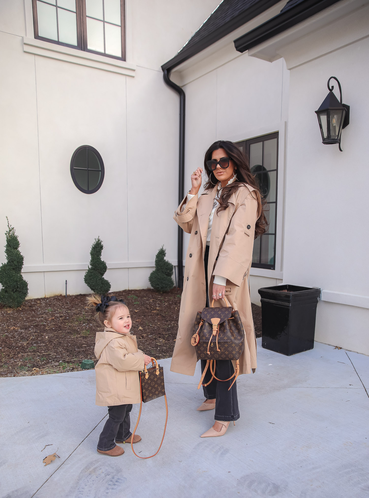 Express spring fashion 2021, emily gemma, mommy daughter matching outfits pinterest, baby fashion blogger, express trench coat 2021 | Mommy and Me Outfits by popular US fashion blog, The Sweetest Thing: image of Emily Gemma and her daughter Sophie standing outside and wearing a Express trench coat, Express flare jeans, Express gold hoop earrings, Express ruffle lack mock neck shirt, and holding a Louis Vuitton bags. 