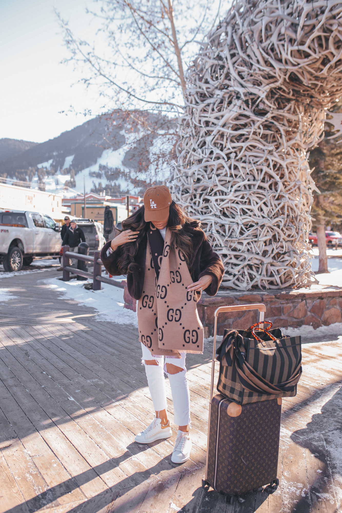 casual travel outfit idea winter 2021, airport fashion outfit idea, cute airport outfit idea winter, fendi tote bag2 |Winter Fashion by popular US fashion blog, The Sweetest Thing: image of Emily Gemma standing outside in downtown Jackson Hole, WY and wearing a Free People top, BB Dakota faux fur jacket, distressed white denim jeans, Gucci scarf, '47 brand cap, Le Specs sunglasses, and standing next to a Louis Vuitton rolling suitcase and holding a Fendi tote bag. 