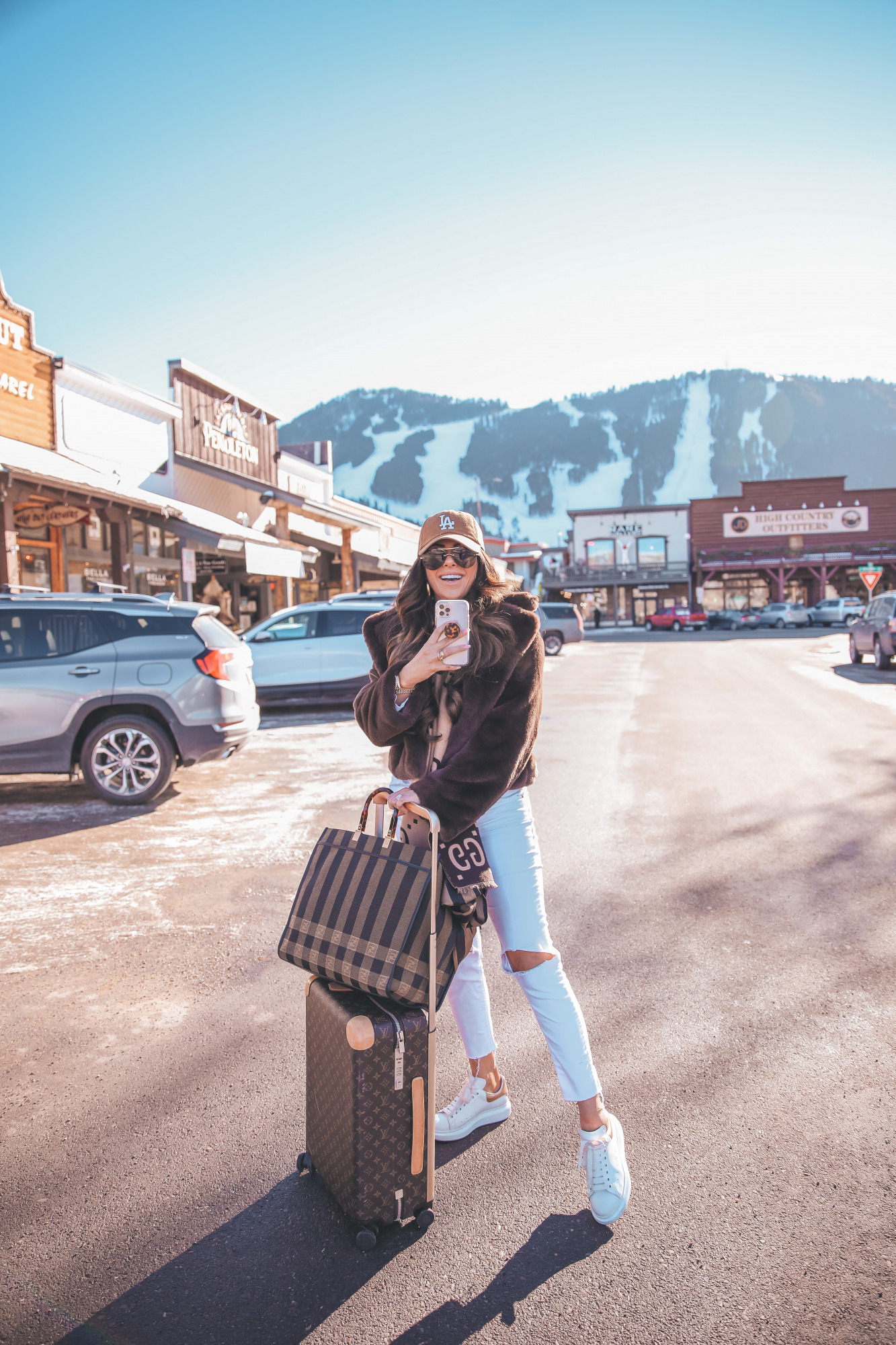 casual travel outfit idea winter 2021, airport fashion outfit idea, cute airport outfit idea winter, fendi tote bag2 |Winter Fashion by popular US fashion blog, The Sweetest Thing: image of Emily Gemma standing outside in downtown Jackson Hole, WY and wearing a Free People top, BB Dakota faux fur jacket, distressed white denim jeans, Gucci scarf, '47 brand cap, Le Specs sunglasses, and standing next to a Louis Vuitton rolling suitcase and holding a Fendi tote bag. 