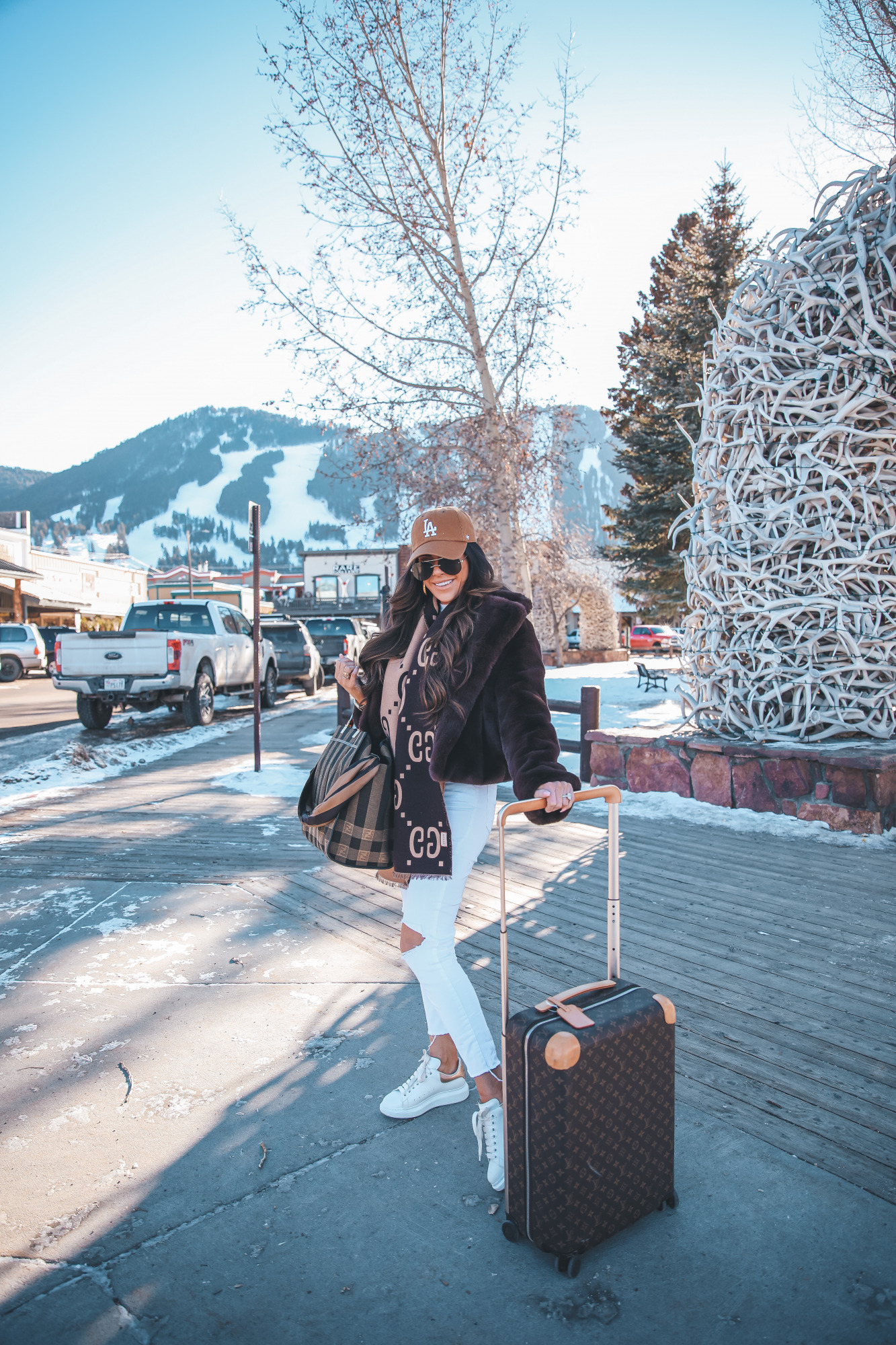 casual travel outfit idea winter 2021, airport fashion outfit idea, cute airport outfit idea winter, fendi tote | Winter Fashion by popular US fashion blog, The Sweetest Thing: image of Emily Gemma standing by the antler arch in Jackson Hole, WY and wearing a Free People top, BB Dakota faux fur jacket, distressed white denim jeans, Gucci scarf, '47 brand cap, Le Specs sunglasses, and standing next to a Louis Vuitton rolling suitcase and holding a Fendi tote bag. 