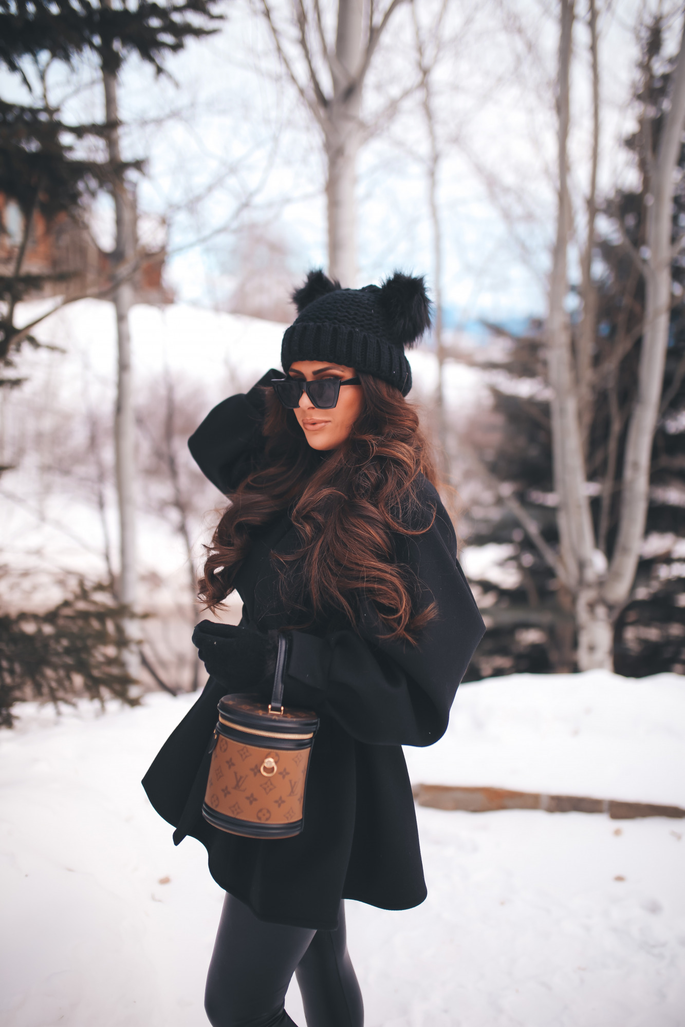 hair by chrissy extensions brunette balayage, emily gemma, chanel necklaceJPG |Dressing Up by popular US fashion blog, The Sweetest Thing: image of Emily Gemma standing outside in the snow and wearing a Good American bodysuit, commando leggings, Chanel boots, Amazon sunglasses, Storets puff sleeve coat, Tasha double pom beanie, and holding a Louis Vuitton bag. 