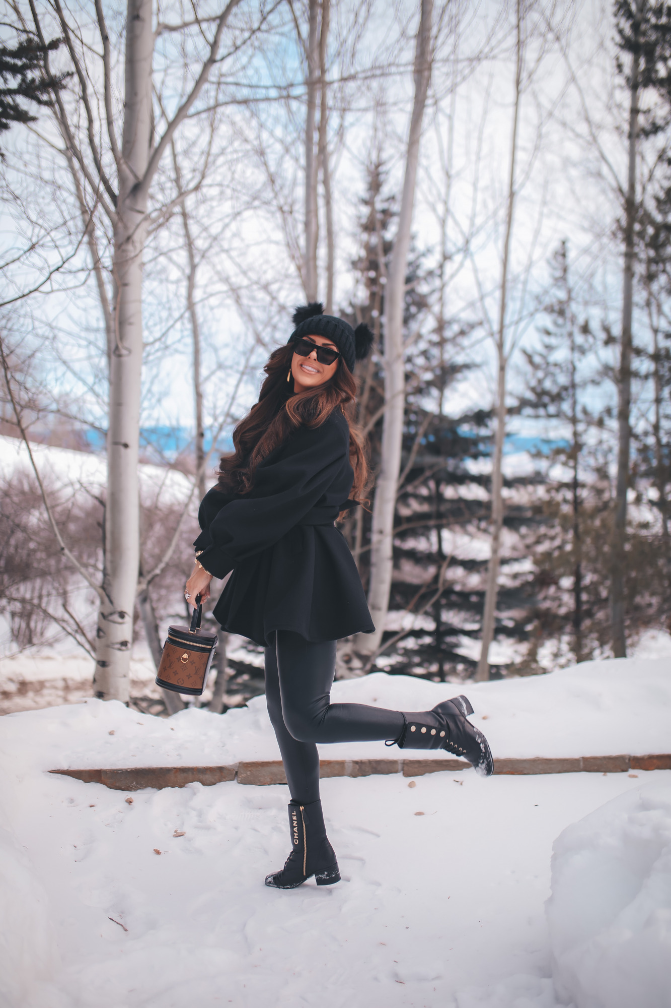 hair by chrissy extensions brunette balayage, emily gemma, pinterest winter outfit idea 2021. | Dressing Up by popular US fashion blog, The Sweetest Thing: image of Emily Gemma standing outside in the snow and wearing a Good American bodysuit, commando leggings, Chanel boots, Amazon sunglasses, Storets puff sleeve coat, Tasha double pom beanie, and holding a Louis Vuitton bag. 