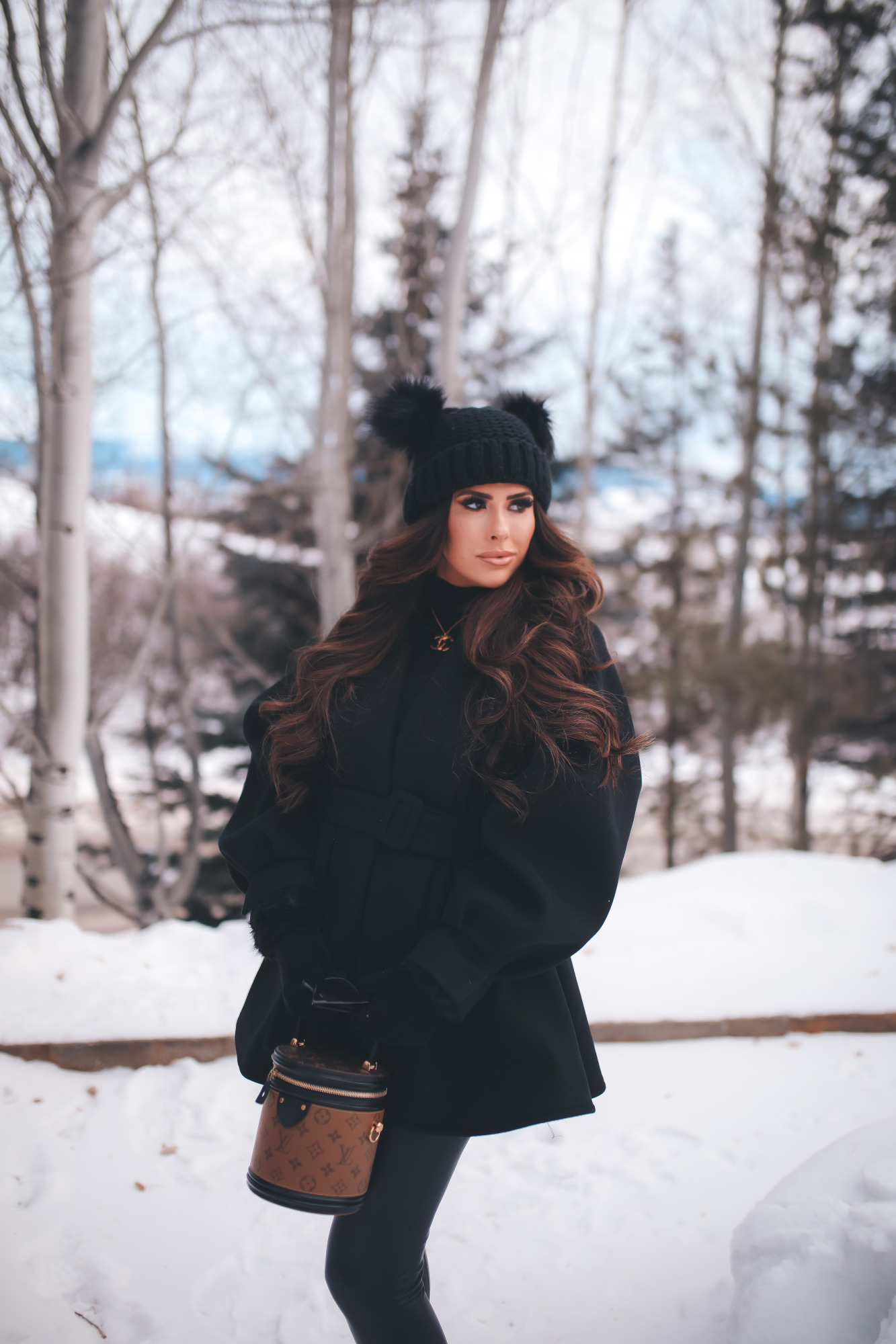 hair by chrissy extensions brunette balayage, emily gemma1, pinterest winter outfit idea 2021 |Dressing Up by popular US fashion blog, The Sweetest Thing: image of Emily Gemma standing outside in the snow and wearing a Good American bodysuit, commando leggings, Chanel boots, Amazon sunglasses, Storets puff sleeve coat, Tasha double pom beanie, and holding a Louis Vuitton bag.