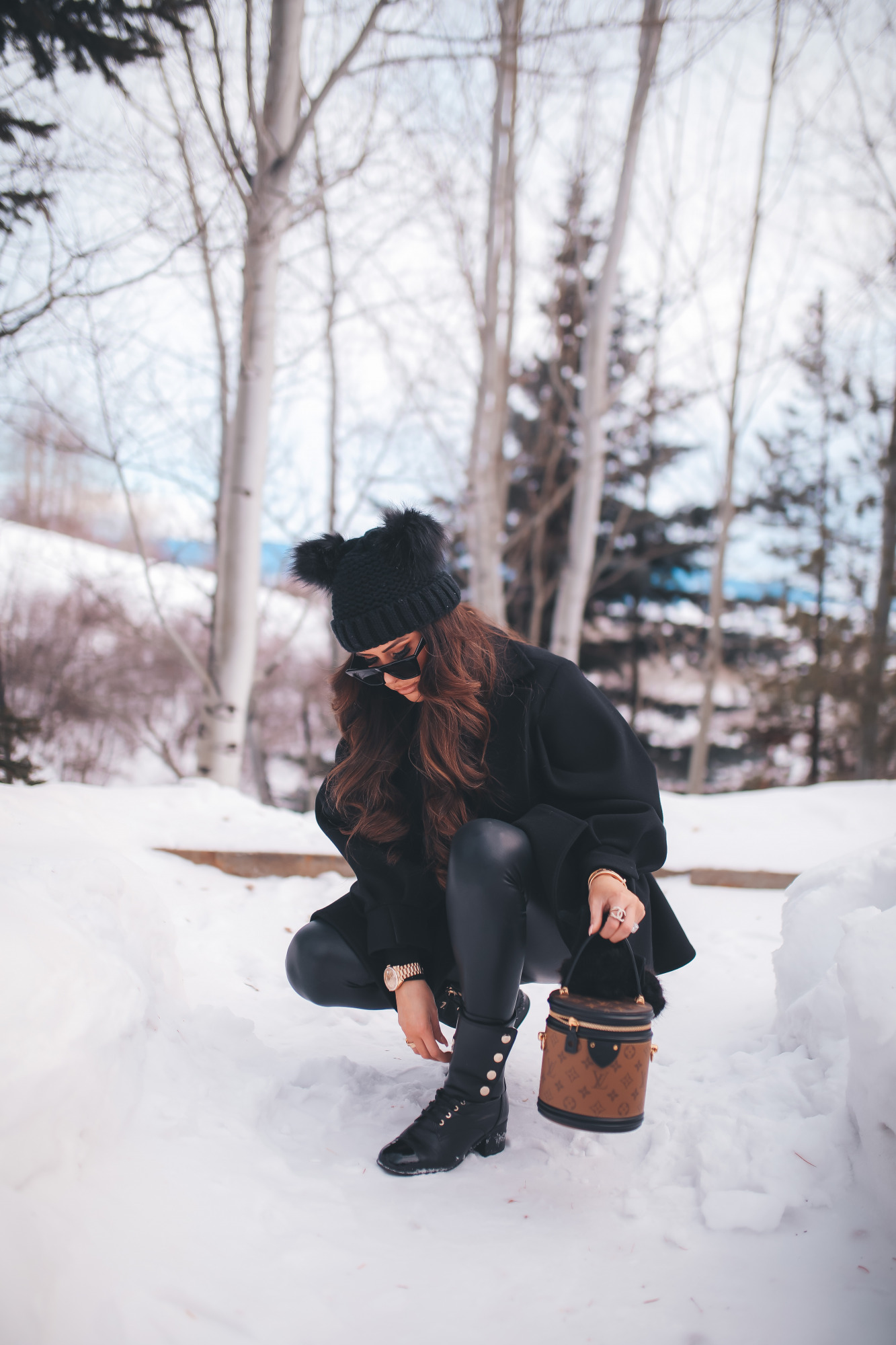 louis vuitton cannes bag, gold 40mm rolex day date, chanel ring, cartier bracelet, emily gemma2 |Dressing Up by popular US fashion blog, The Sweetest Thing: image of Emily Gemma standing outside in the snow and wearing a Good American bodysuit, commando leggings, Chanel boots, Amazon sunglasses, Storets puff sleeve coat, Tasha double pom beanie, and holding a Louis Vuitton bag. 