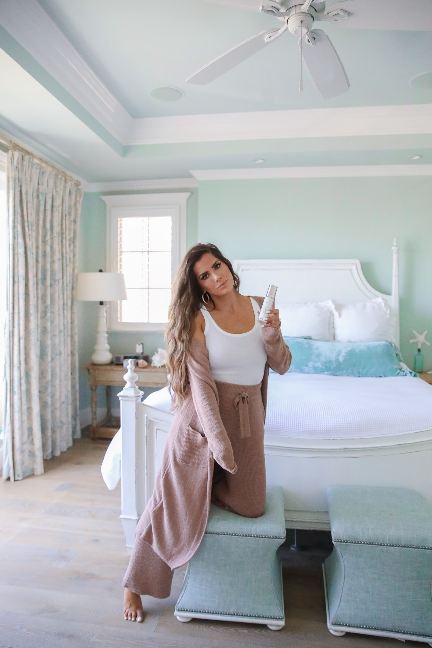 Top Picks by popular US life and style blog, The Sweetest Thing: image of a woman wearing a mauve sweater set and white tank while kneeling on a blue stool in front of a bed with a white wood bed frame. 