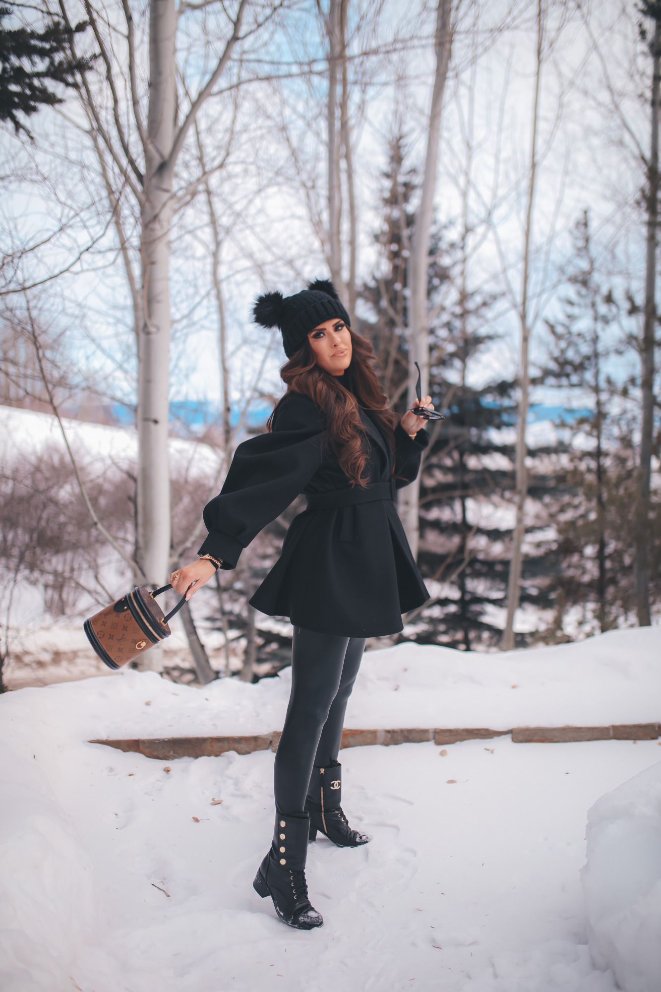 winter fashion outfit ideas pinterest, chanel combat boots, emily gemma, pinterest winter outfit idea 2021.2 |Dressing Up by popular US fashion blog, The Sweetest Thing: image of Emily Gemma standing outside in the snow and wearing a Good American bodysuit, commando leggings, Chanel boots, Amazon sunglasses, Storets puff sleeve coat, Tasha double pom beanie, and holding a Louis Vuitton bag. 