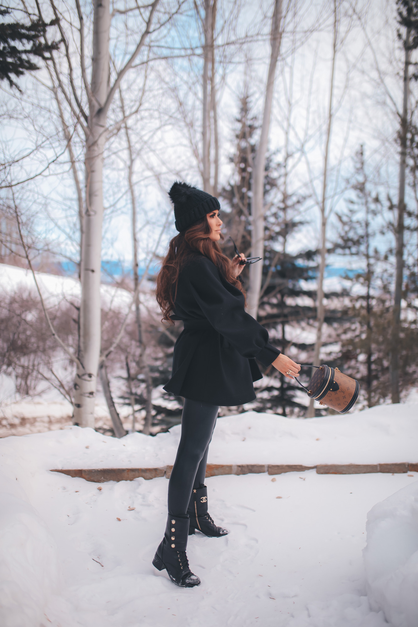 winter fashion outfit ideas pinterest, chanel combat boots, emily gemma, pinterest winter outfit idea 202122 |Dressing Up by popular US fashion blog, The Sweetest Thing: image of Emily Gemma standing outside in the snow and wearing a Good American bodysuit, commando leggings, Chanel boots, Amazon sunglasses, Storets puff sleeve coat, Tasha double pom beanie, and holding a Louis Vuitton bag. 