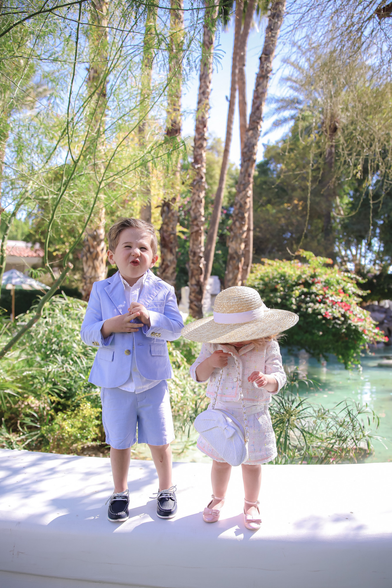 Spring Outfit Ideas for Kids by popular US fashion blog, The Sweetest Thing: image of a young boy and girl standing outside next to a landscaped pond and wearing a Pink Boucle Jacket, Pink Boucle Shorts, Pink Bow Flats, White Purse, Straw Sun Hat, Seersucker Blazer, Seersucker Shorts, White Button-down, and Leather Boat Shoes.