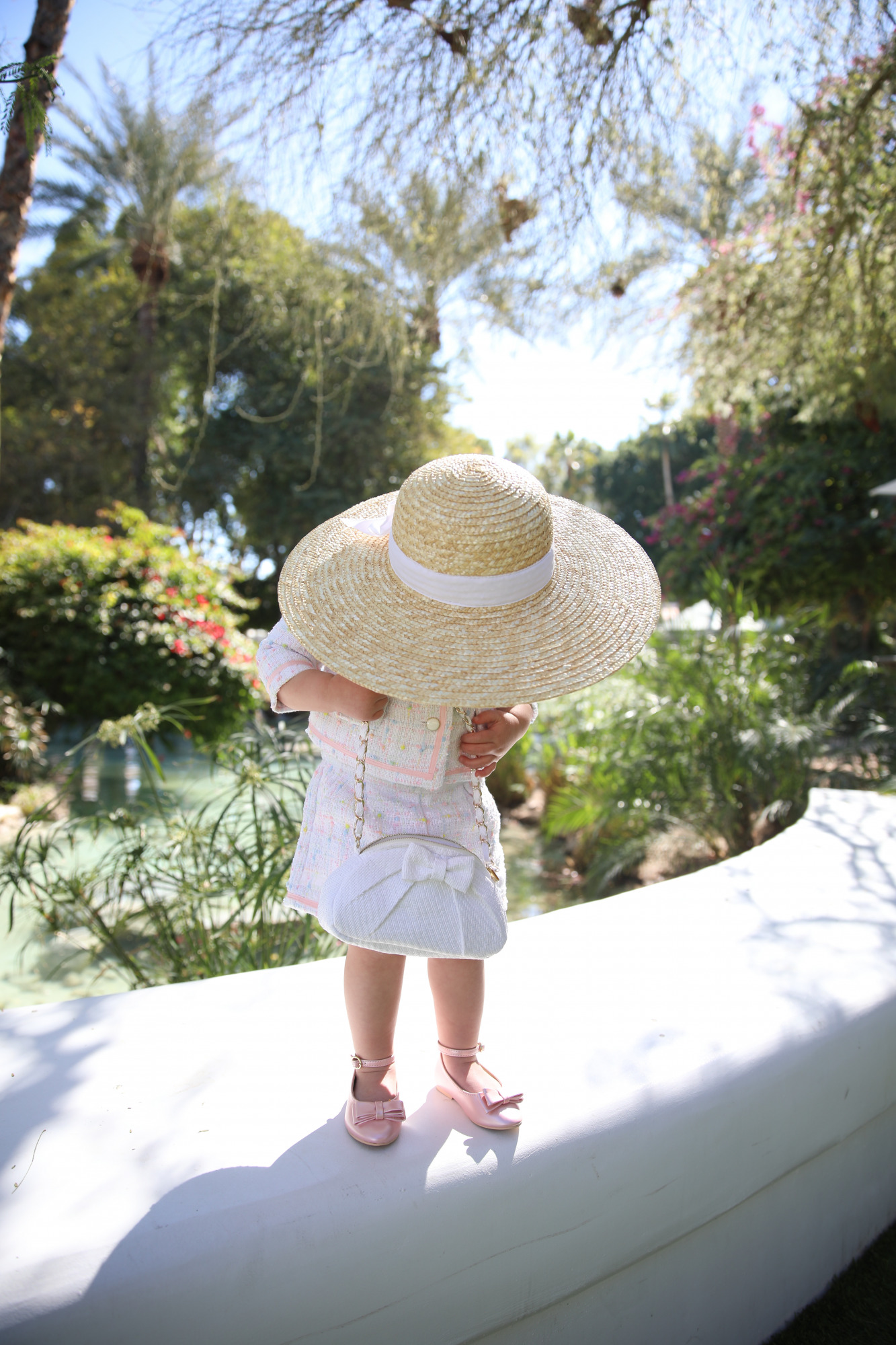 janie and jack easter kids outfits 2021, kids easter outfit ideas toddler, emily gemma, scottsdale fashion blog4 |Spring Outfit Ideas for Kids by popular US fashion blog, The Sweetest Thing: image of a young girl standing outside next to a landscaped pond and wearing a Pink Boucle Jacket, Pink Boucle Shorts, Pink Bow Flats, White Purse, and Straw Sun Hat.