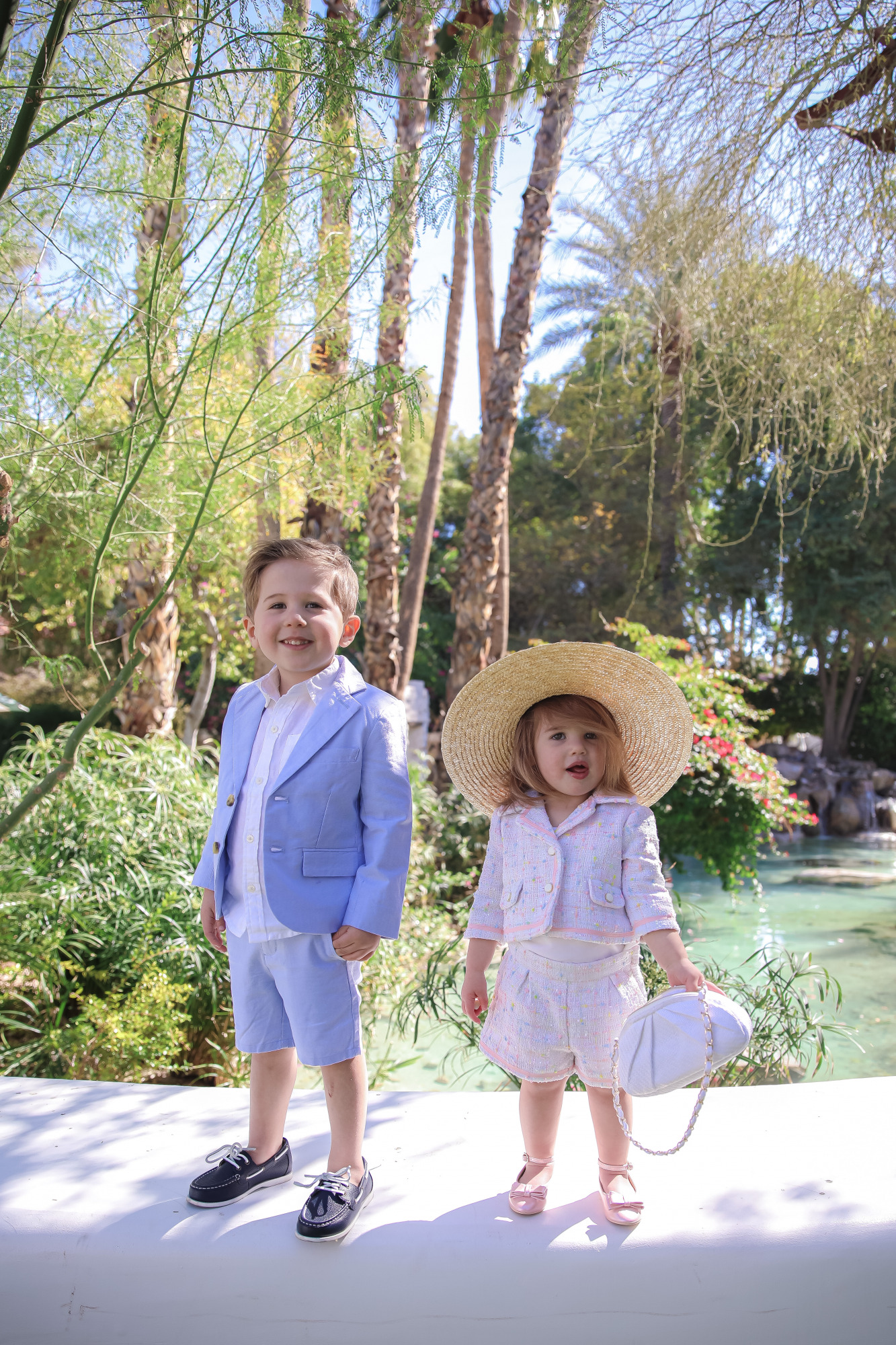 janie and jack easter kids outfits 2021, kids easter outfit ideas toddler, emily gemma, scottsdale fashion blog4 |Spring Outfit Ideas for Kids by popular US fashion blog, The Sweetest Thing: image of a young boy and girl standing outside next to a landscaped pond and wearing a Pink Boucle Jacket, Pink Boucle Shorts, Pink Bow Flats, White Purse, Straw Sun Hat, Seersucker Blazer, Seersucker Shorts, White Button-down, and Leather Boat Shoes.