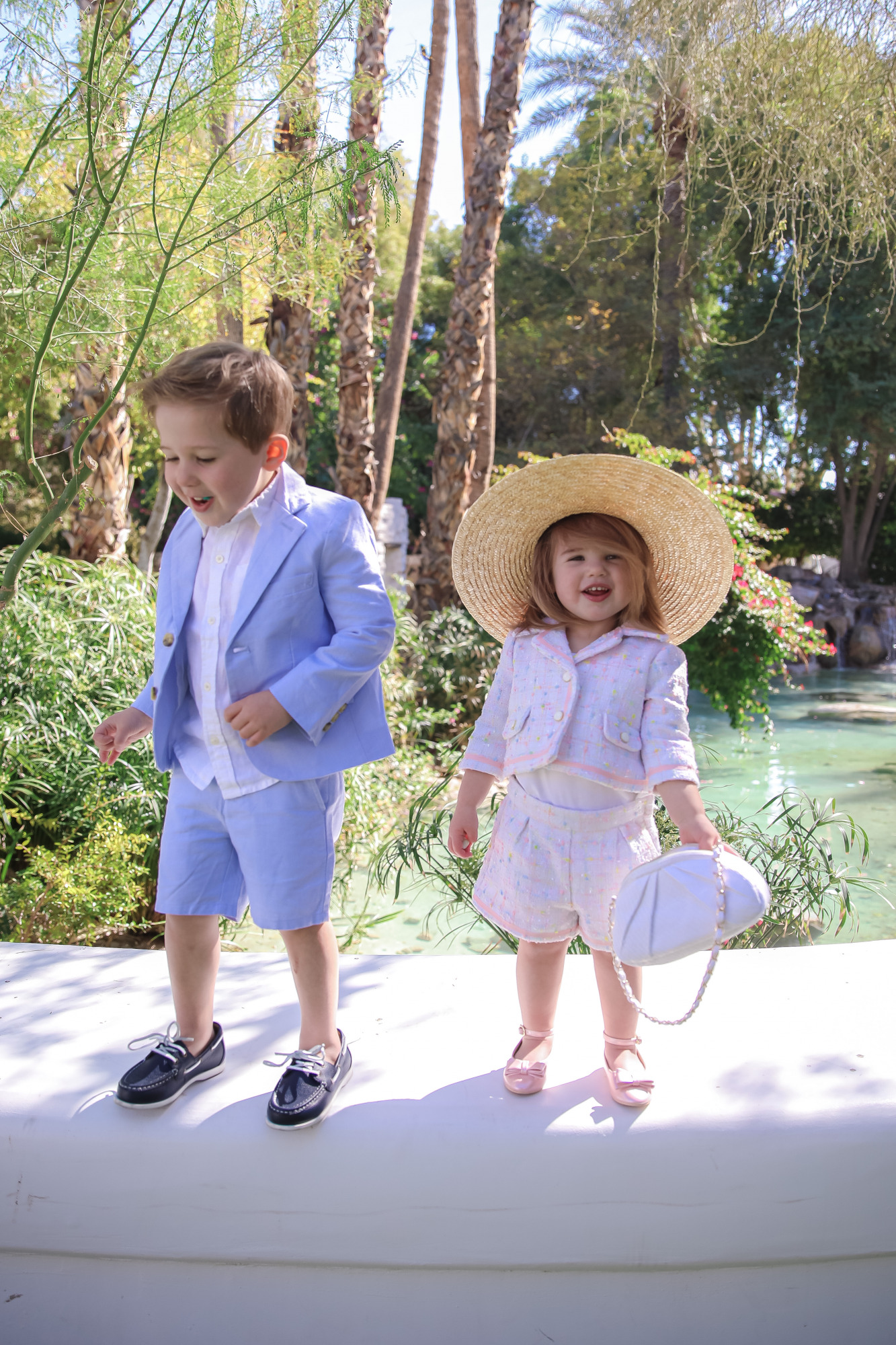 janie and jack easter kids outfits 2021, kids easter outfit ideas toddler, emily gemma, scottsdale fashion blog4 |Spring Outfit Ideas for Kids by popular US fashion blog, The Sweetest Thing: image of a young boy and girl standing outside next to a landscaped pond and wearing a Pink Boucle Jacket, Pink Boucle Shorts, Pink Bow Flats, White Purse, Straw Sun Hat, Seersucker Blazer, Seersucker Shorts, White Button-down, and Leather Boat Shoes.