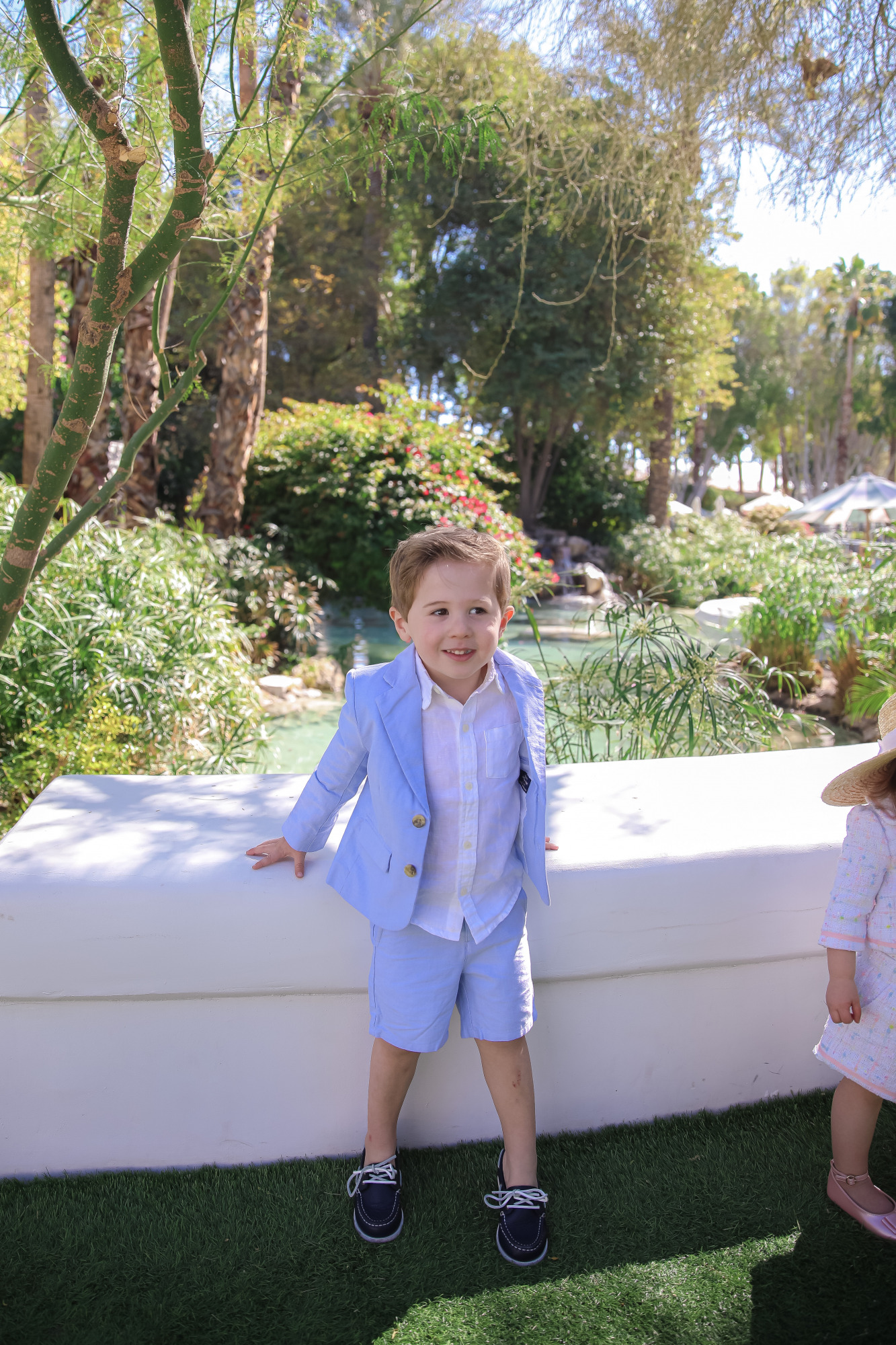 janie and jack easter kids outfits 2021, kids easter outfit ideas toddler, emily gemma, scottsdale fashion blog9 |Spring Outfit Ideas for Kids by popular US fashion blog, The Sweetest Thing: image of a young boy and girl standing outside next to a landscaped pond and wearing a Pink Boucle Jacket, Pink Boucle Shorts, Pink Bow Flats, White Purse, Straw Sun Hat, Seersucker Blazer, Seersucker Shorts, White Button-down, and Leather Boat Shoes.