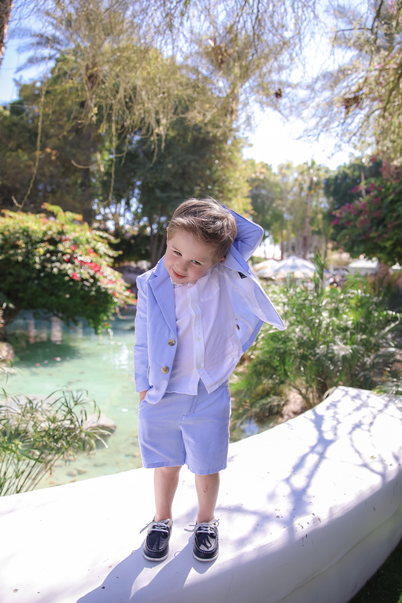 janie and jack easter kids outfits 2021, kids easter outfit ideas toddler, toddler boy seersucker suit |Spring Outfit Ideas for Kids by popular US fashion blog, The Sweetest Thing: image of a young boy standing outside next to a landscaped pond and wearing a Seersucker Blazer, Seersucker Shorts, White Button-down, and Leather Boat Shoes.