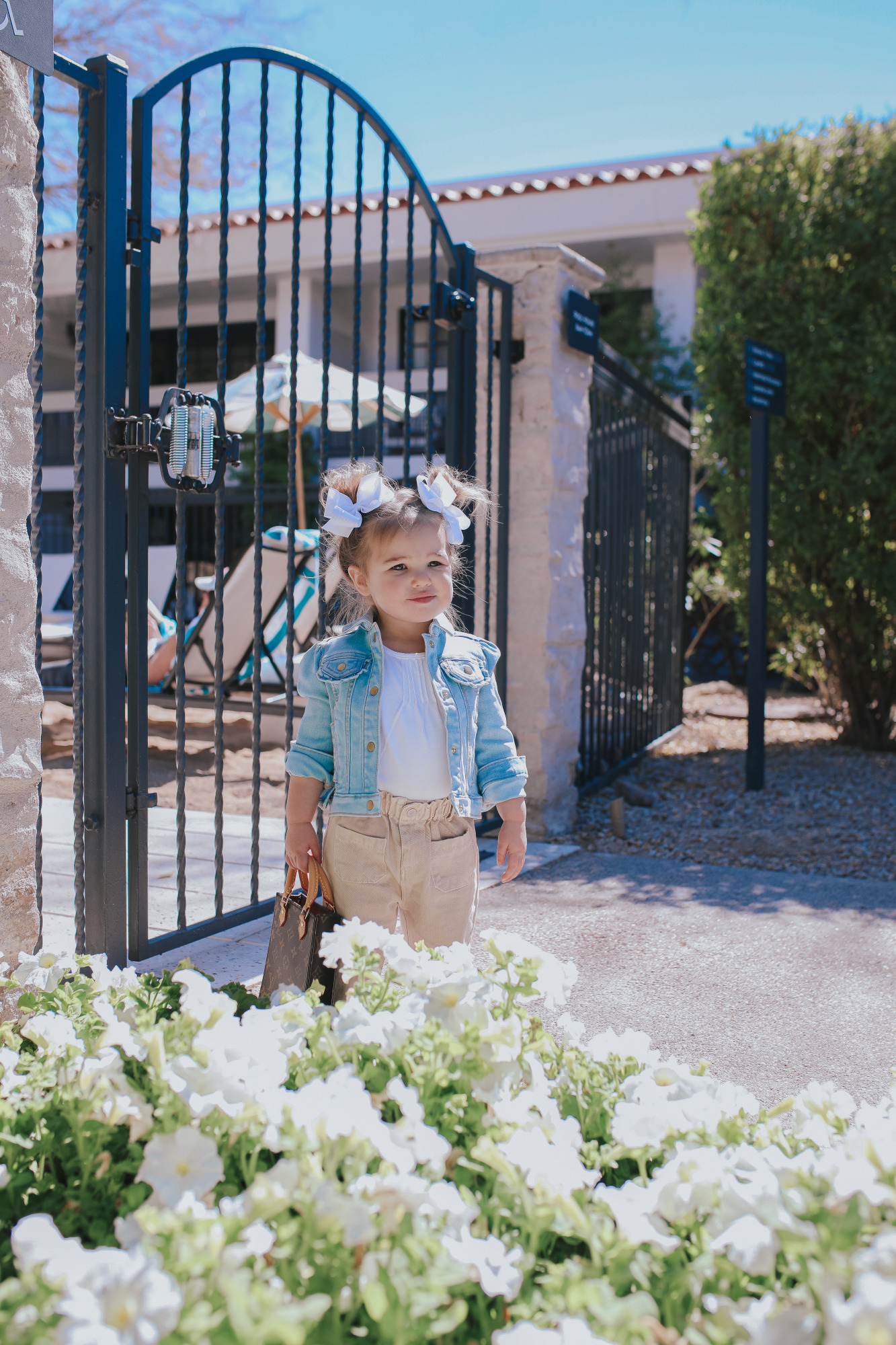 Mom Jeans by popular US fashion blog, The Sweetest Thing: image of a little girl standing by a wrought iron gate in Scottsdale, Arizona and wearing two white hair bows, jean jacket, white shirt, tan pants and holding a mini Louis Vuitton purse. 
