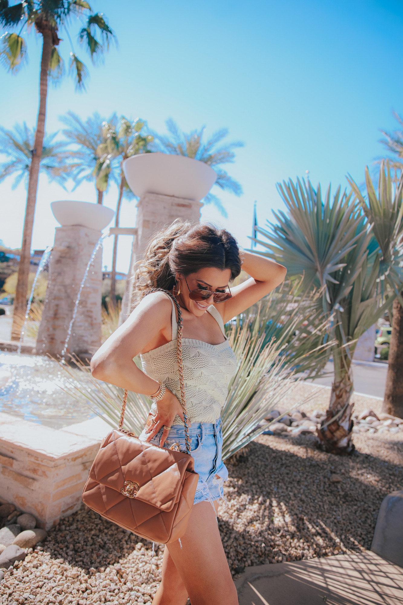 Best distressed high waisted denim shorts summer 2021, tan brown chanel 19 flap bag medium emily gemma0 |High Waisted Denim Shorts by popular US fashion blog, The Sweetest Thing: image of Emily Gemma standing outside and wearing a knit tank top, high waisted denim shorts, woven cross strap sandals, sunglasses and holding a Chanel purse. 