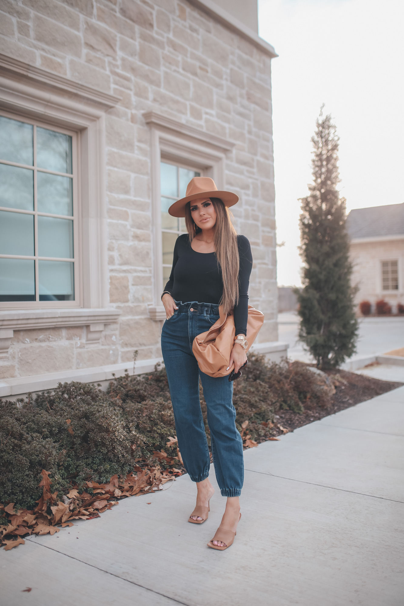 express spring fashion 2021, bottega clutch dupe, denim high waisted joggers, emily gemma15 | Body Contour by popular US fashion blog, The Sweetest Thing: image of a Emily Gemma wearing an Express black bodysuit, Express high waisted joggers, tan heel slide sandals, tan felt fedora, and holding a Bottega clutch dupe. 